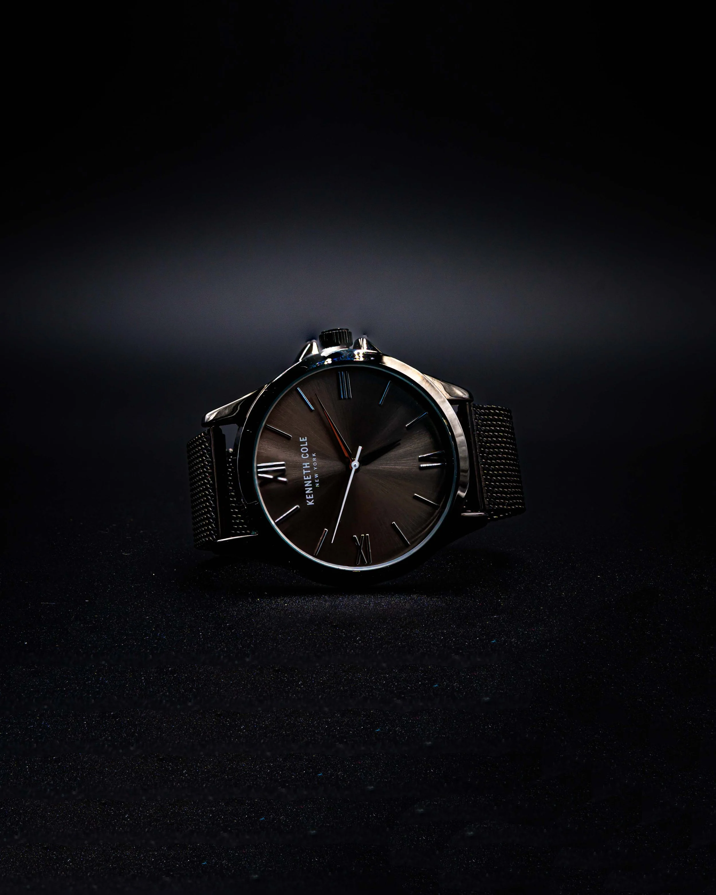 Close-up of a Kenneth Cole wristwatch with a dark face, Roman numerals, and a metal mesh band against a black background.