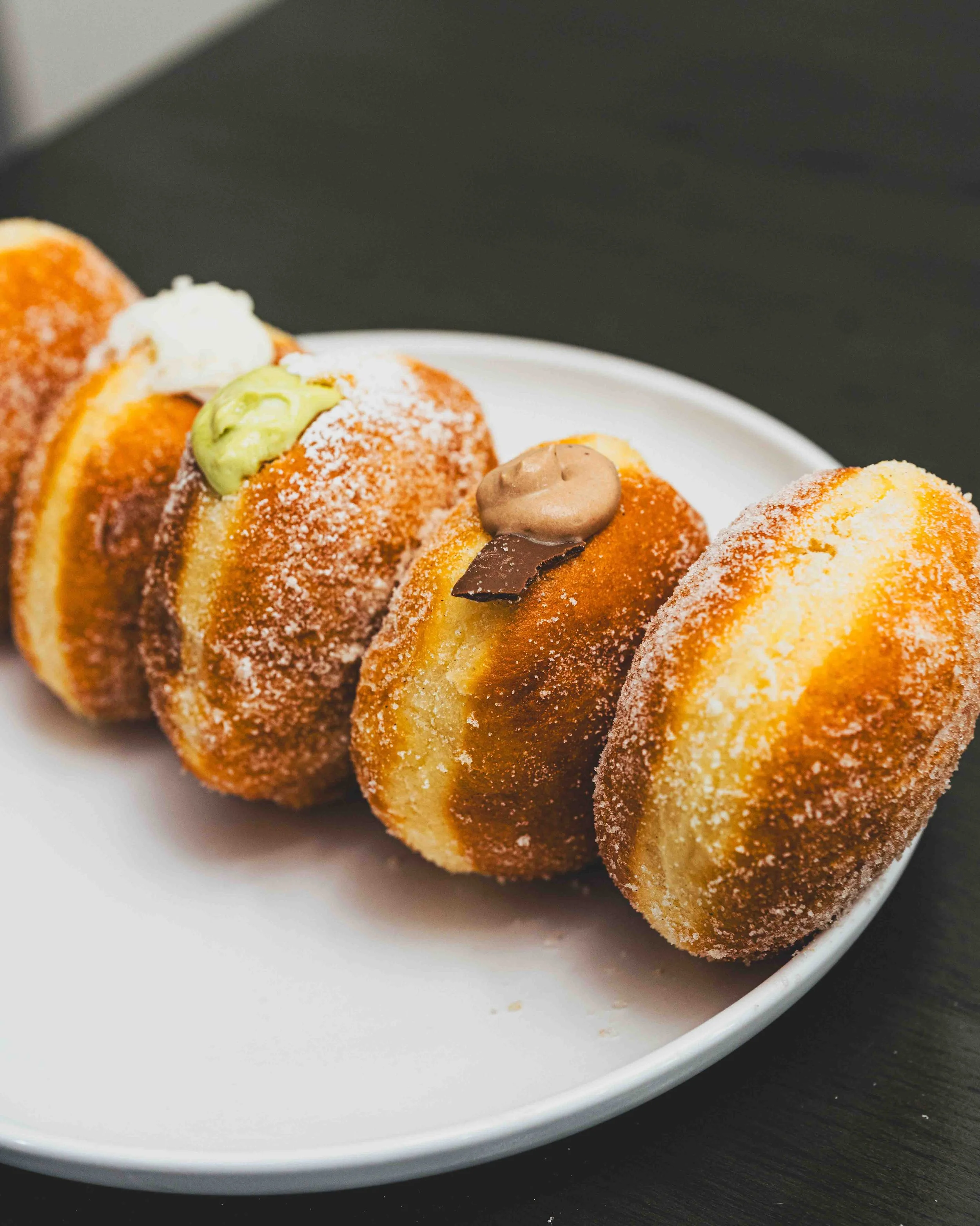 Close-up of three deep-fried dessert balls with sugar, topped with dollops of cream, green, brown, and beige sauces, served on a white plate.