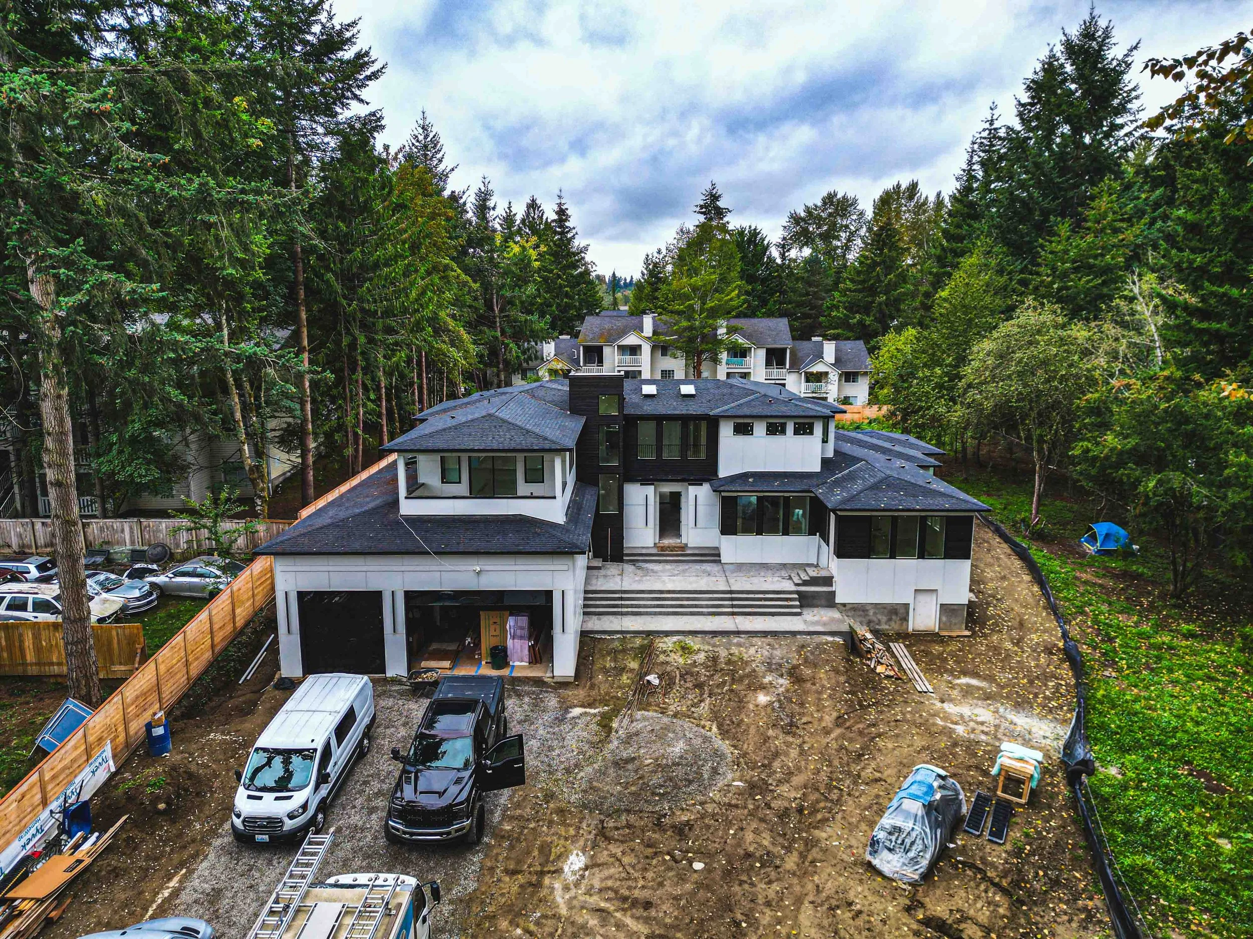 Construction site with multiple modern white and black houses being built among trees, parked cars, and construction materials.