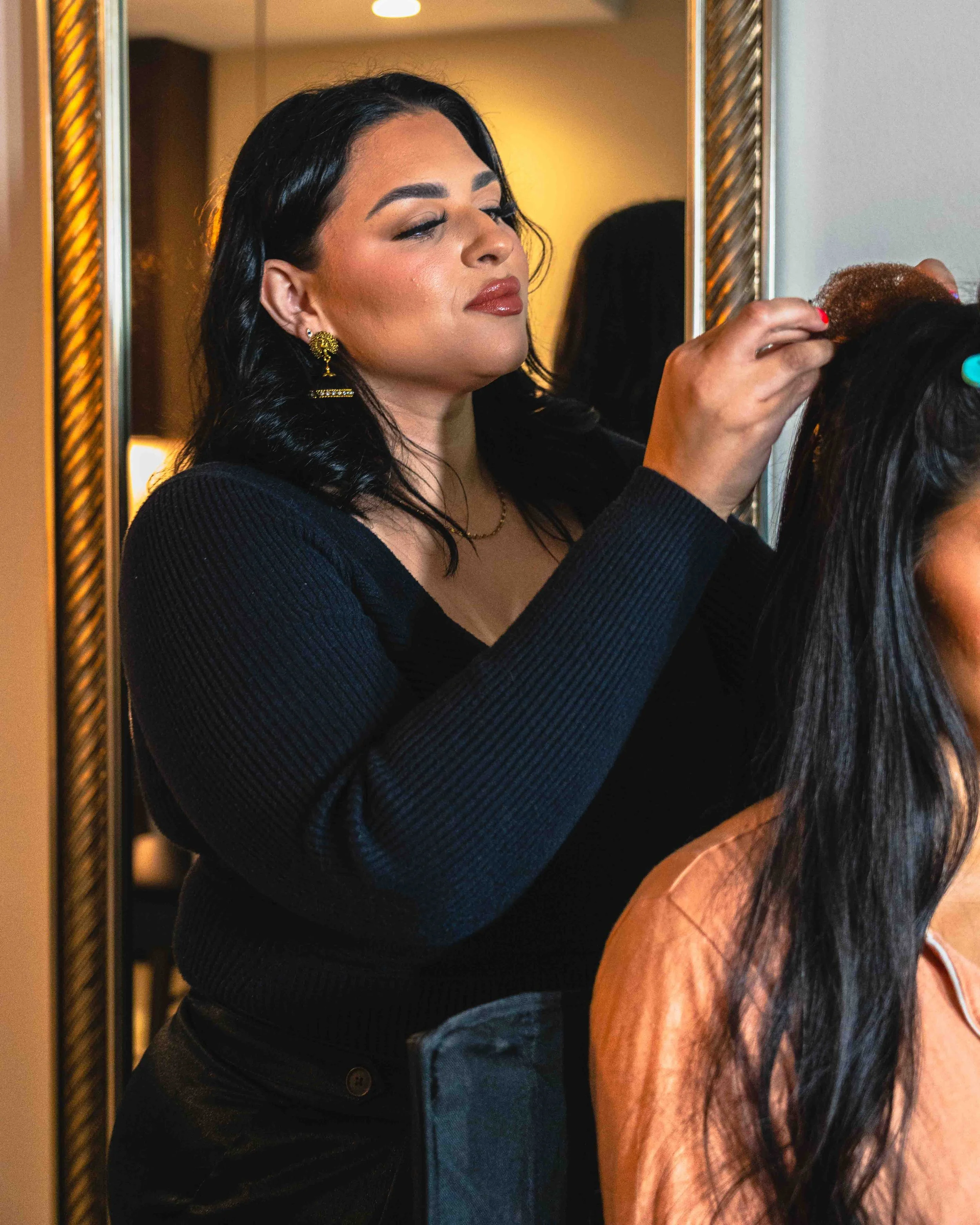 A woman with black hair and gold earrings styling another woman's brown hair in front of a mirror.