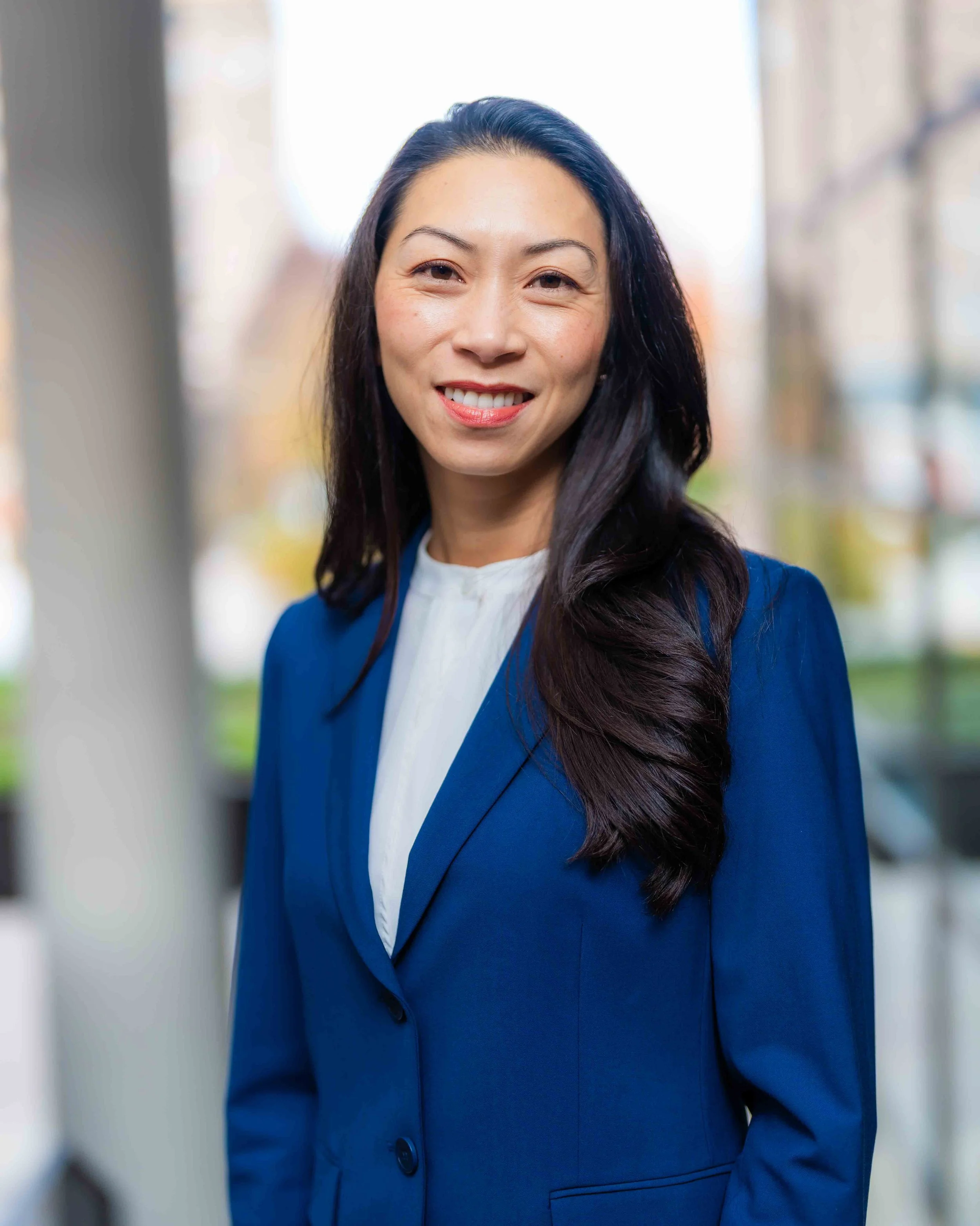 A woman with long dark hair wearing a blue blazer and white shirt, smiling outdoors with blurred trees and buildings in the background.