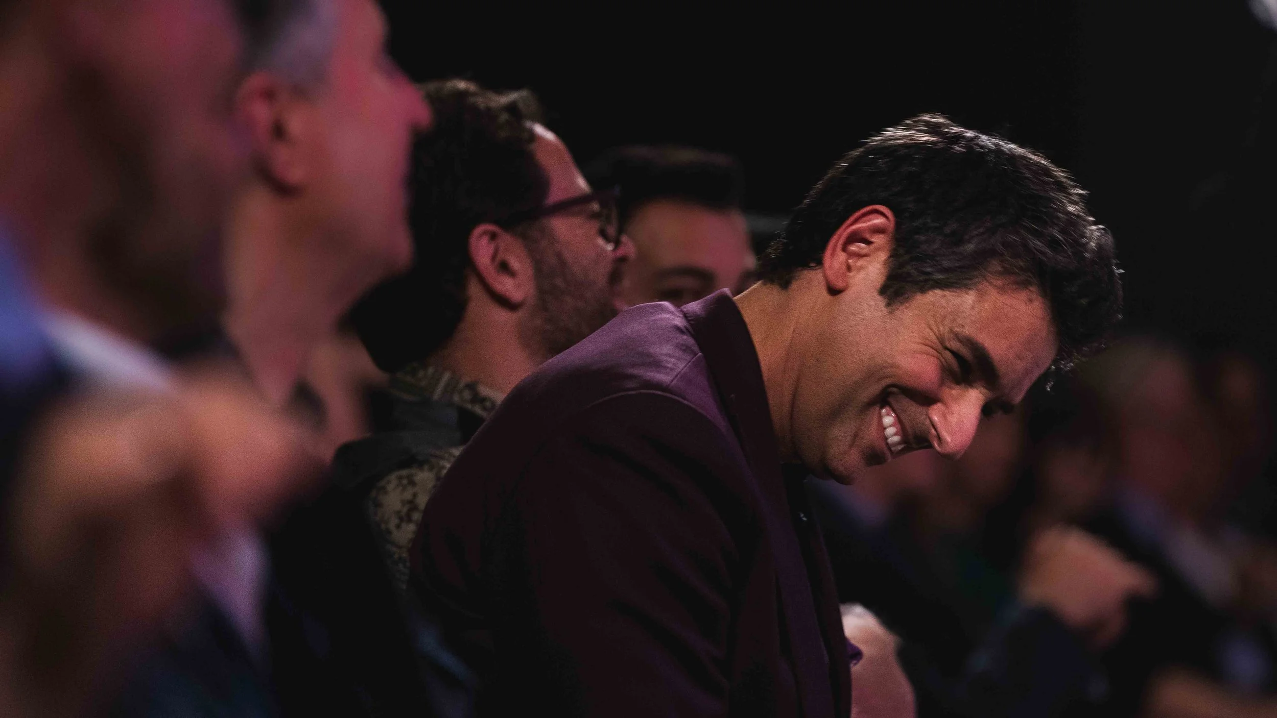 A group of people sitting at an event, smiling and laughing, with a dark background.