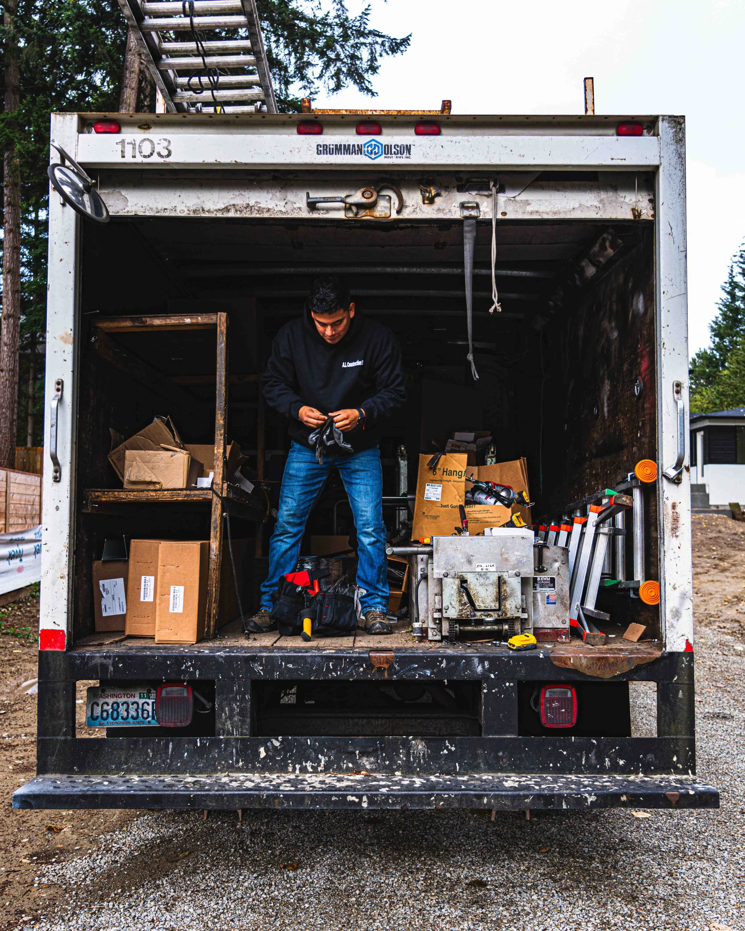 Man standing inside the back of an open delivery truck, organizing tools and boxes, with a ladder on the roof and trees in the background.