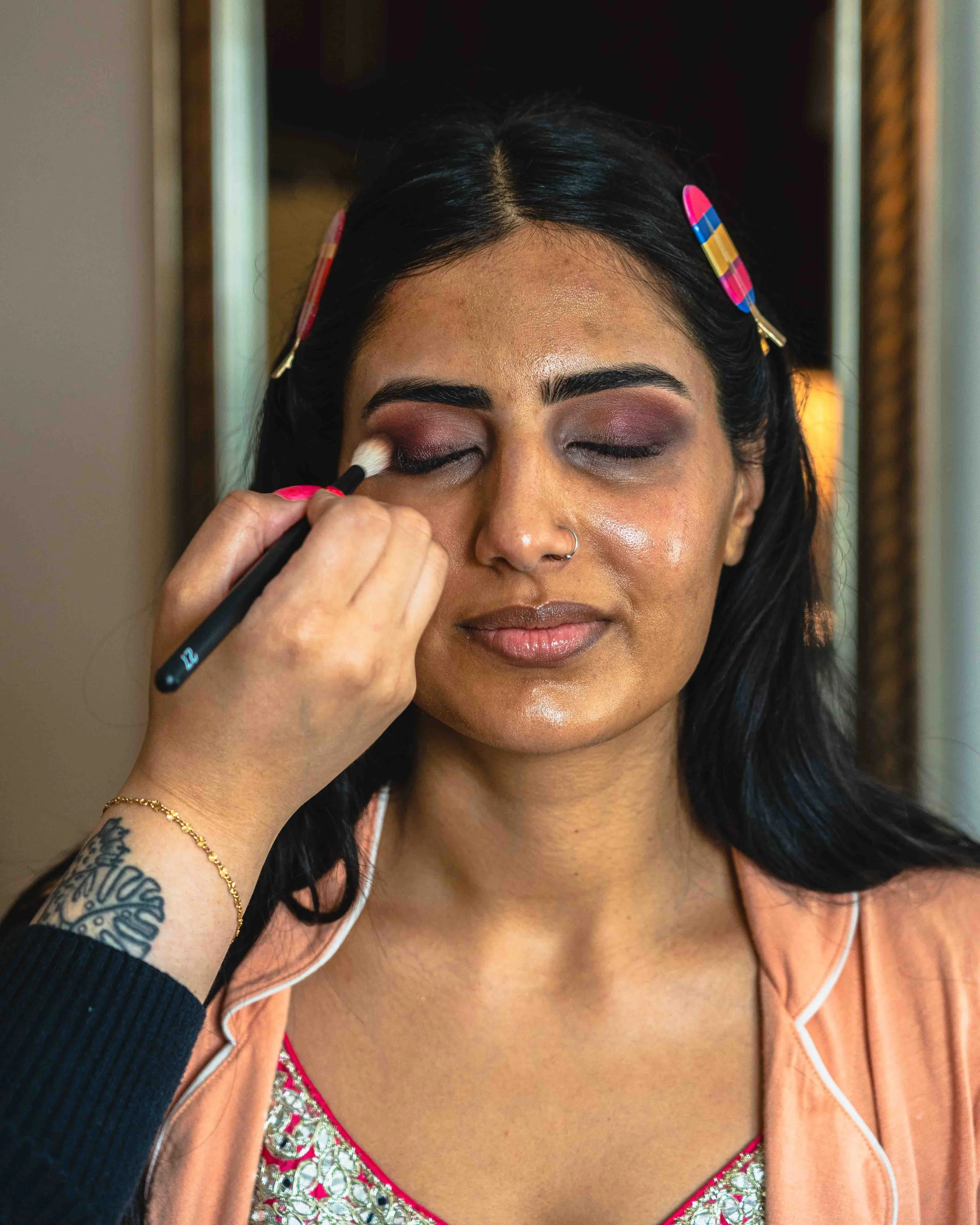 A woman with black hair and brown skin is having makeup applied to her eyelids. She has a nose piercing and is wearing a peach-colored top with white trim, a patterned pink and silver top underneath, and gold jewelry. Her eyes are closed as makeup is