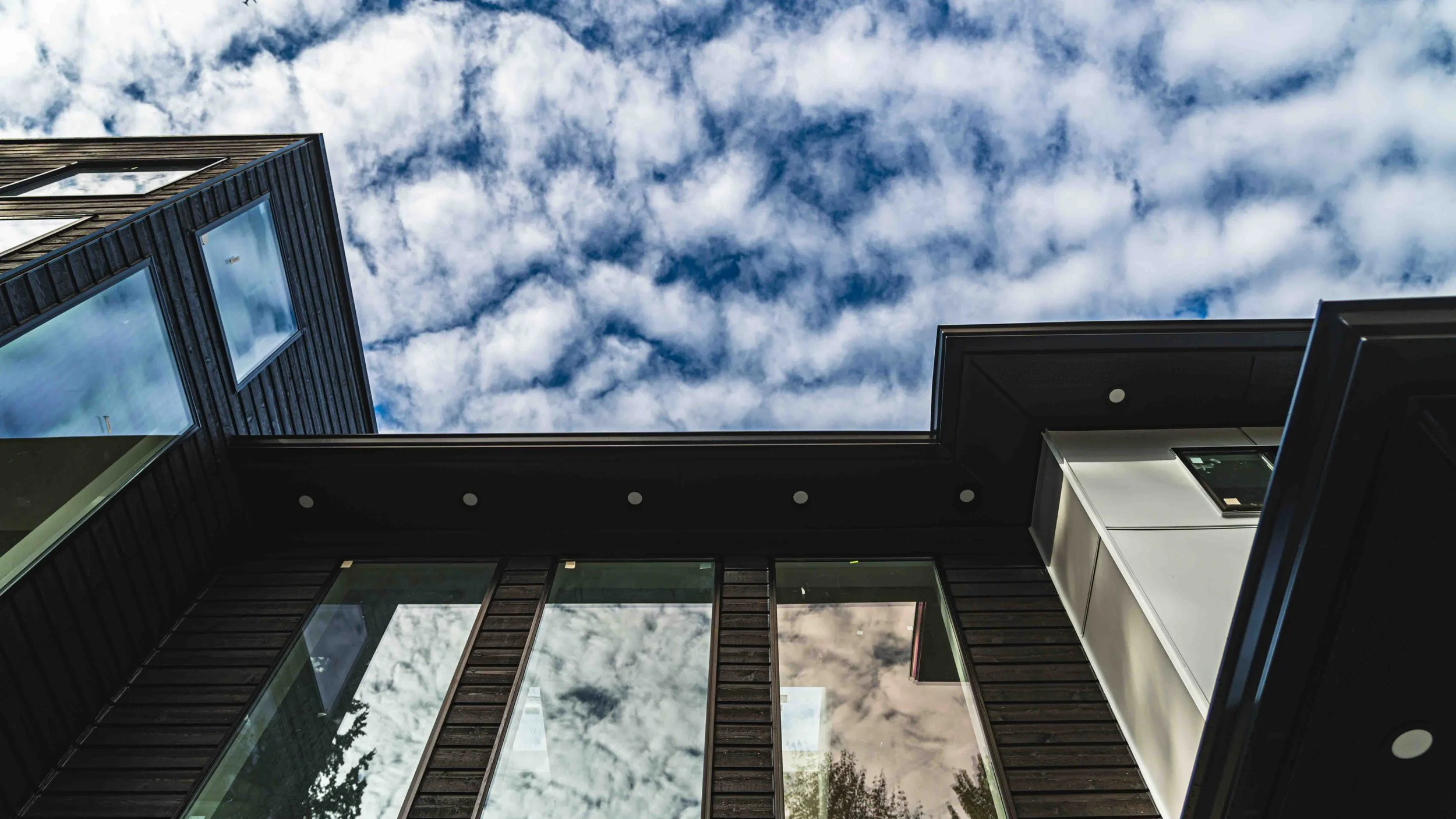 Looking up at modern buildings with large windows and black siding, with a partly cloudy sky overhead.