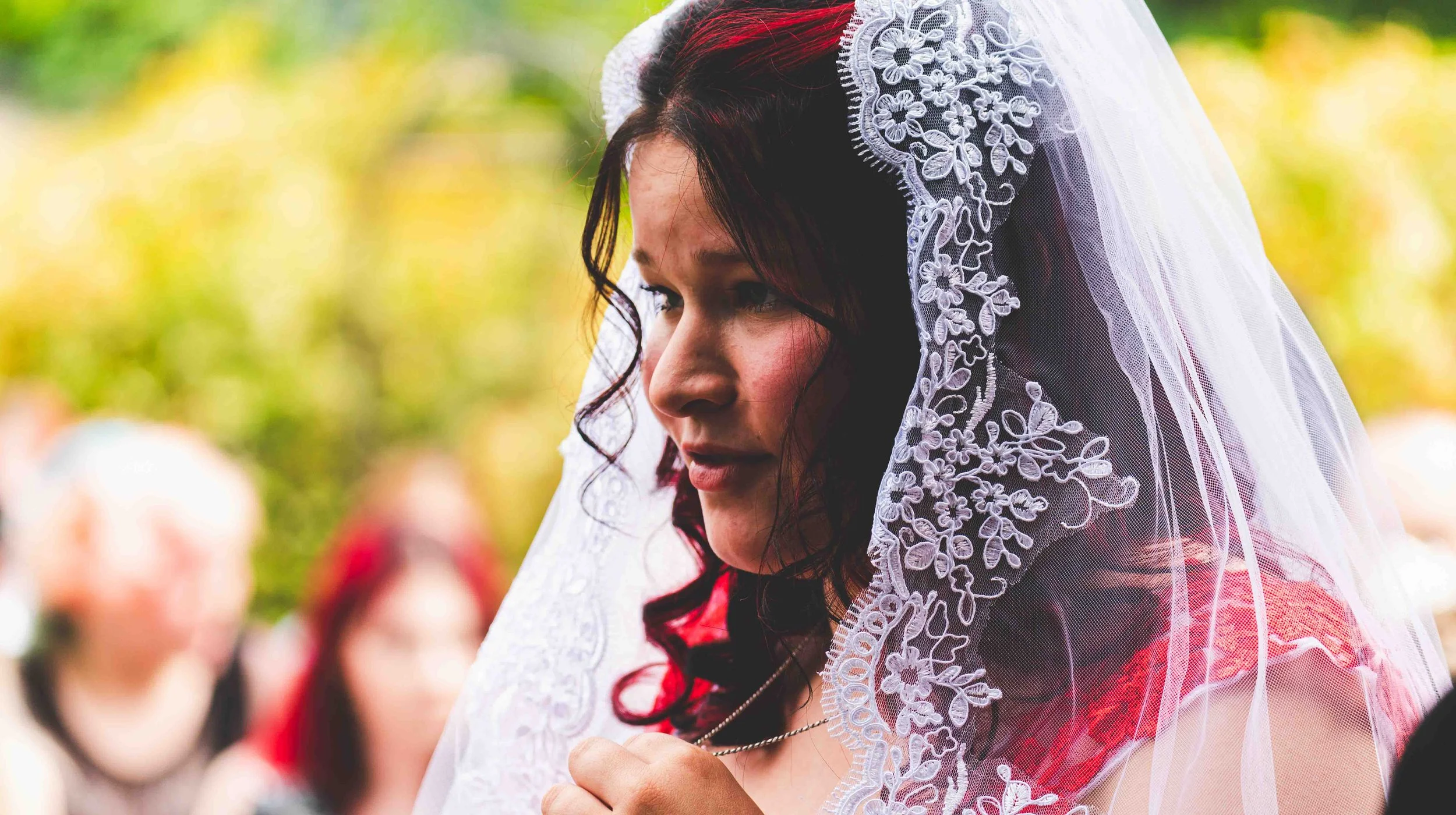 A woman with dark curly hair wearing a lace veil, participating in a wedding ceremony outdoors with a blurred crowd in the background.