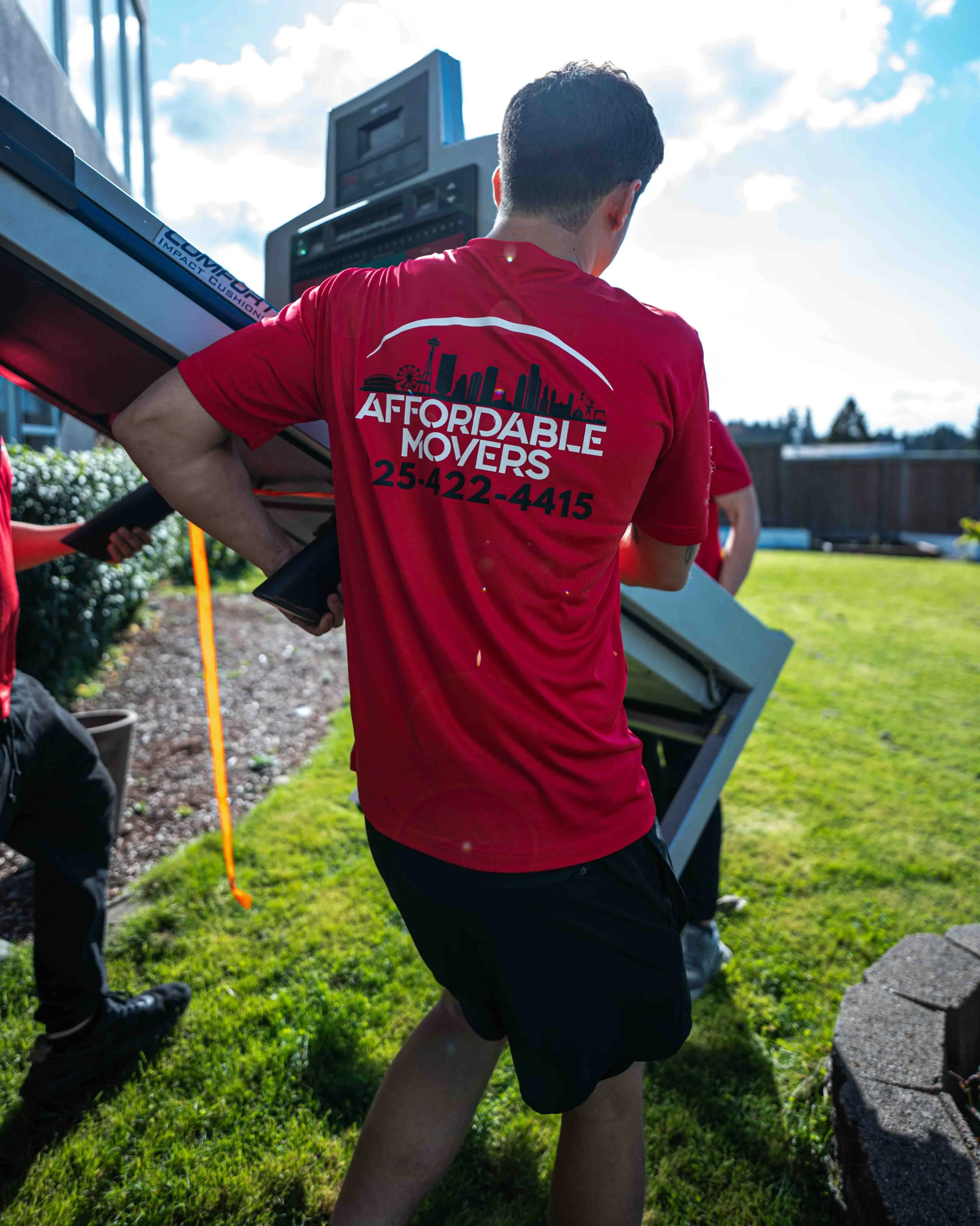 A person wearing a red shirt with a cityscape graphic and the words "Affordable Movers" on the back, carrying a piece of furniture outdoors during daytime. There are other people and greenery in the background.