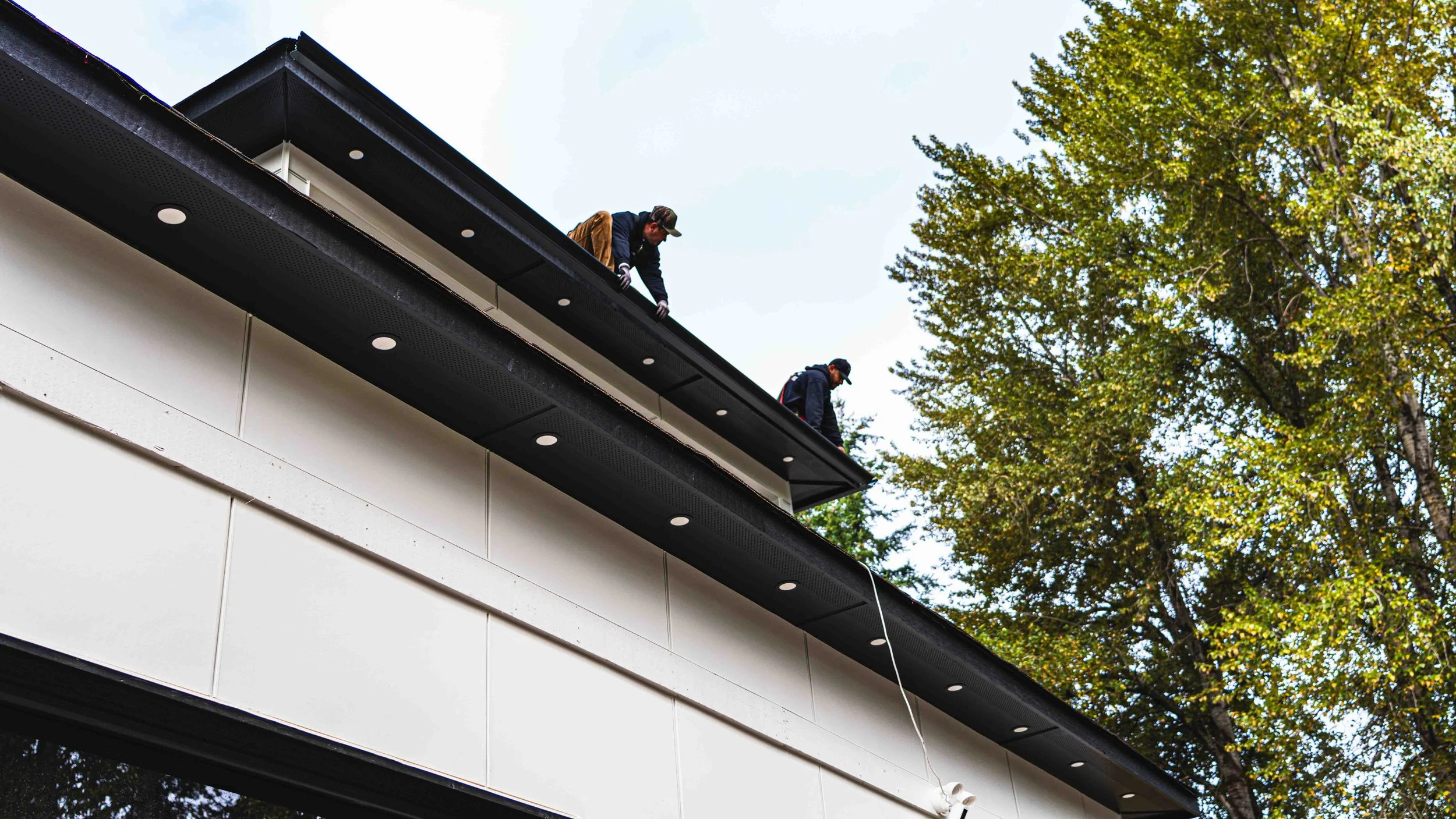 Two workers on a balcony of a modern building, repairing or inspecting the roof, with trees and blue sky in the background.