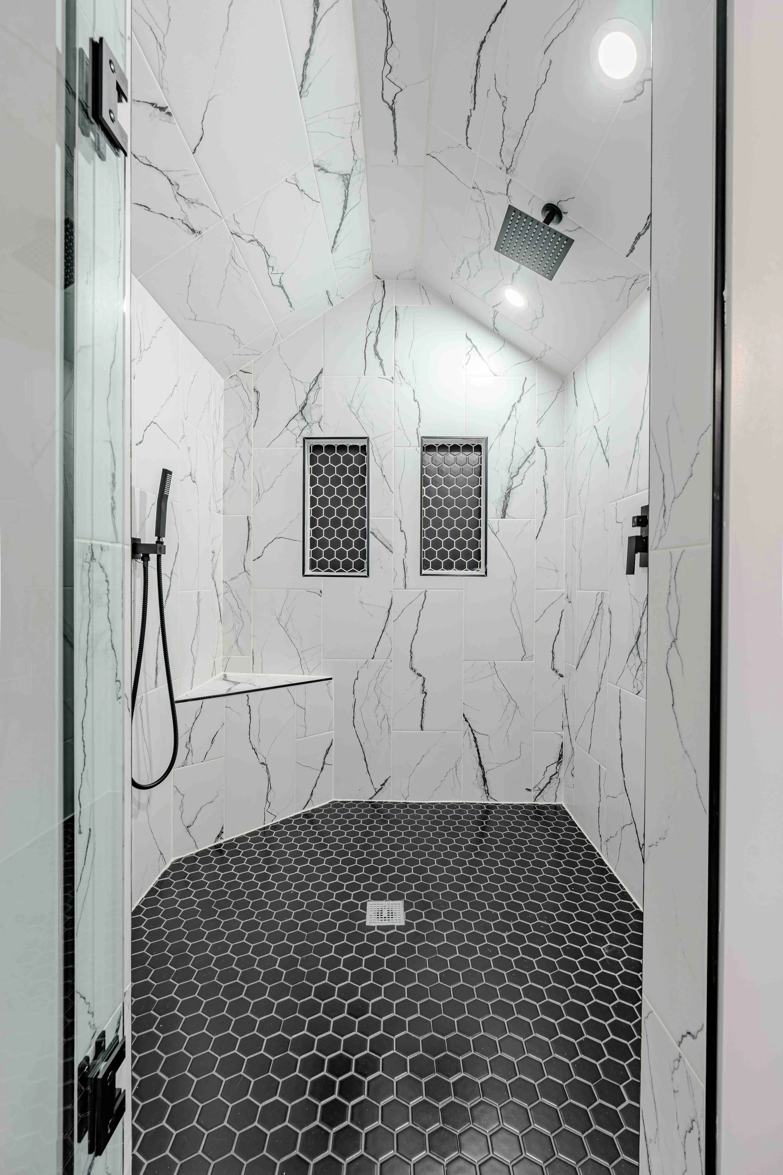 Modern walk-in shower with white marble walls featuring black veins, black hexagonal floor tiles, two black square wall vents, a black showerhead, and a flexible shower hand wand, viewed through a glass door.