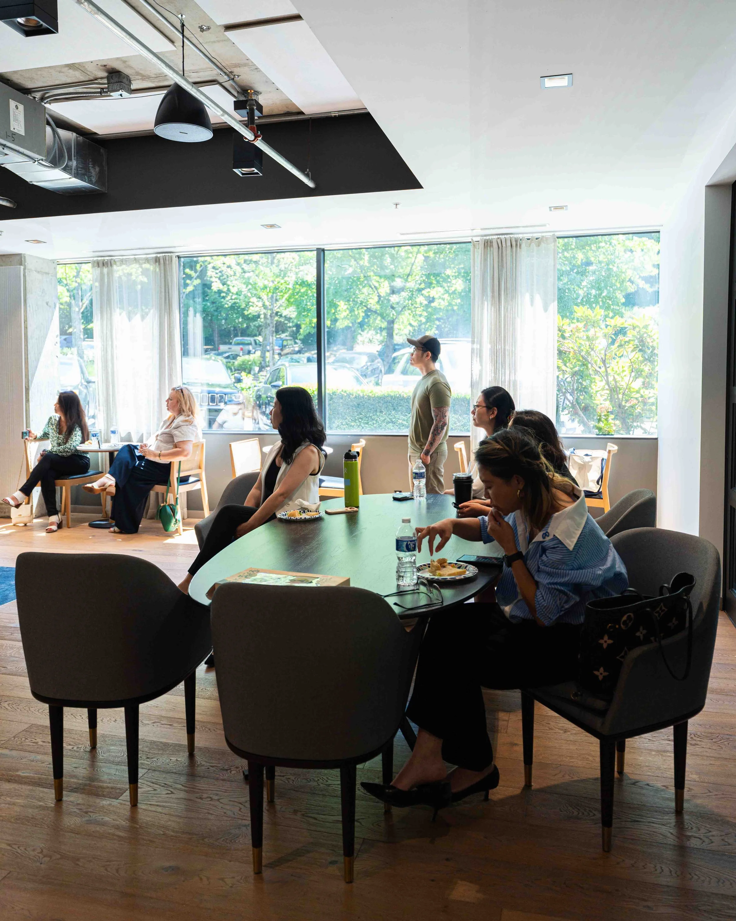 Interior of a modern office or conference room with several women and one man seated or standing, some using phones, others looking forward or to the side, with large windows showing green trees and parked cars outside.