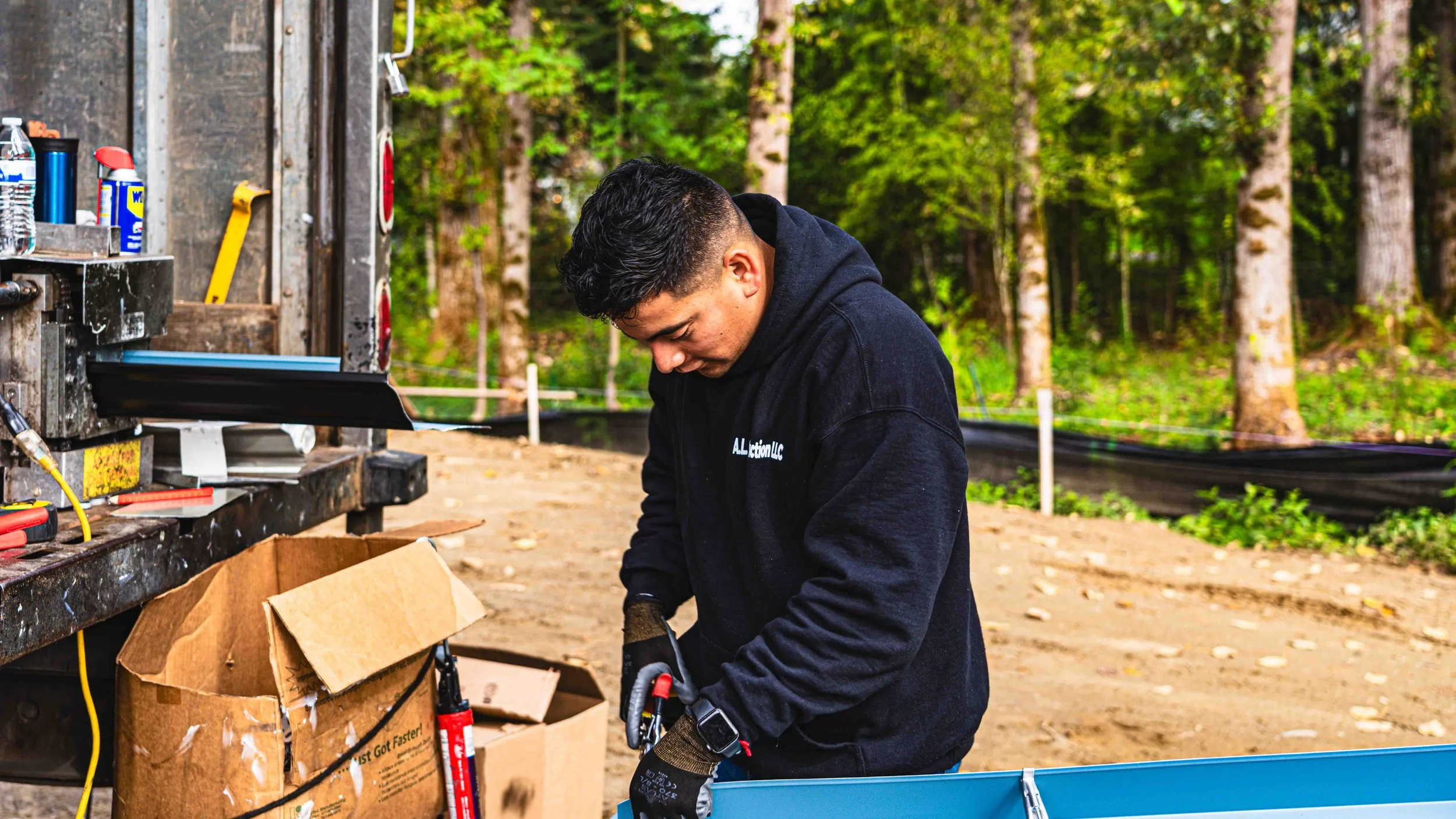 A man working outdoors with trees in the background, wearing a black hoodie, with tools and supplies around him, including a cardboard box and equipment on a workbench.