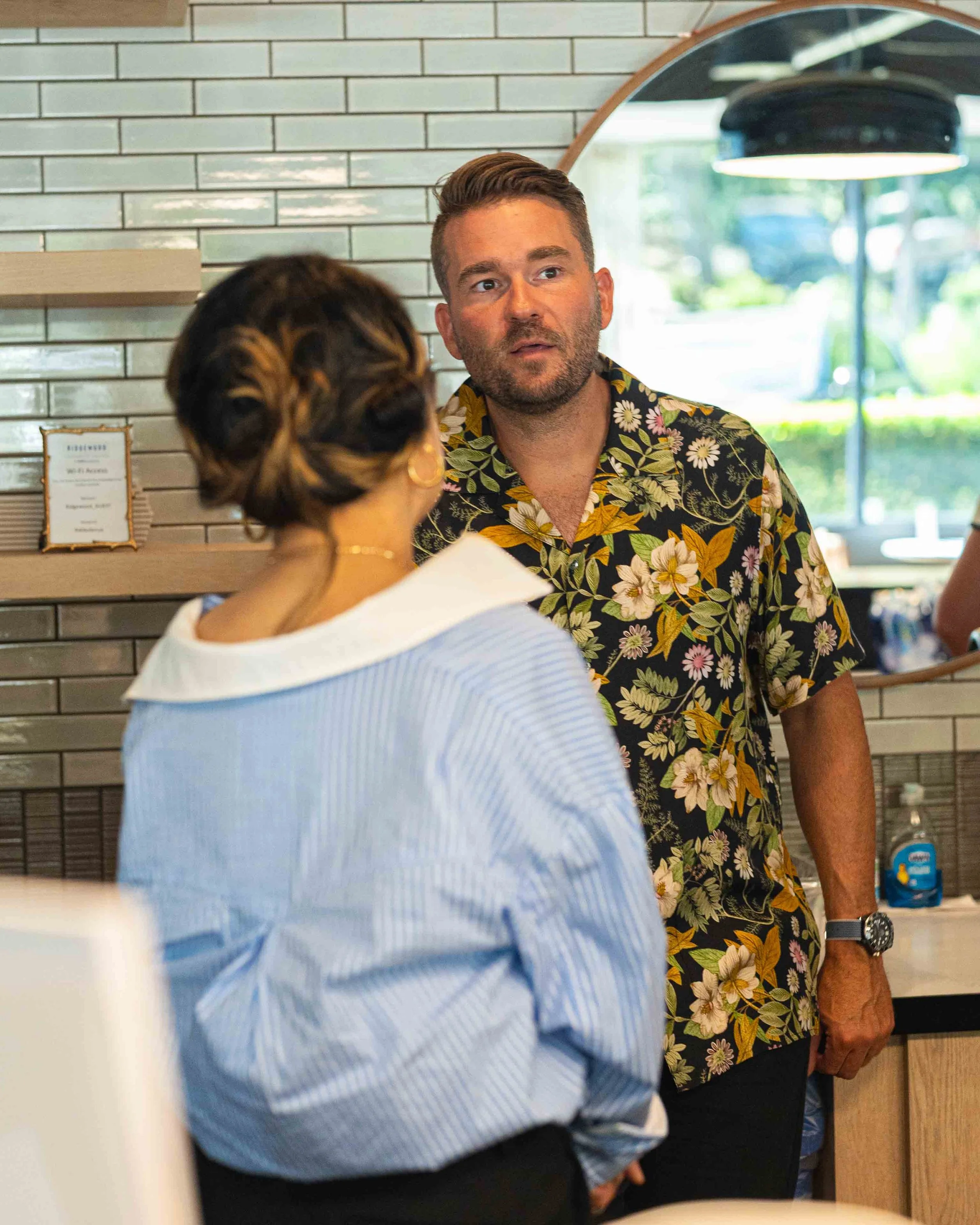 A man wearing a colorful floral shirt and a watch is in a conversation with a woman whose hair is styled in an elegant updo with blond highlights. They are indoors with a modern, tiled wall behind them and a large window showing greenery outside.