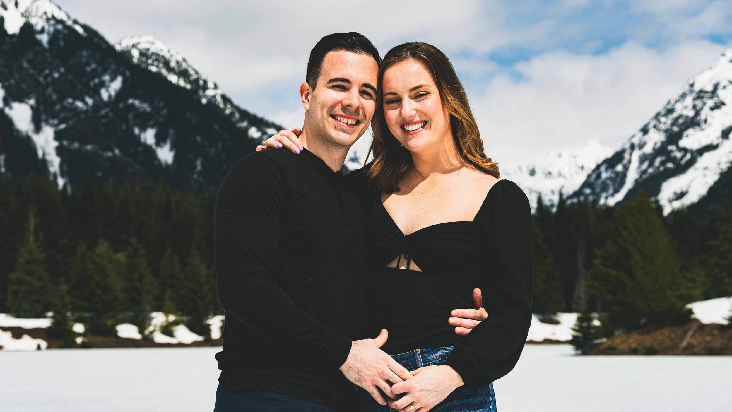 A smiling couple standing close together outdoors with snow-covered mountains and trees in the background.