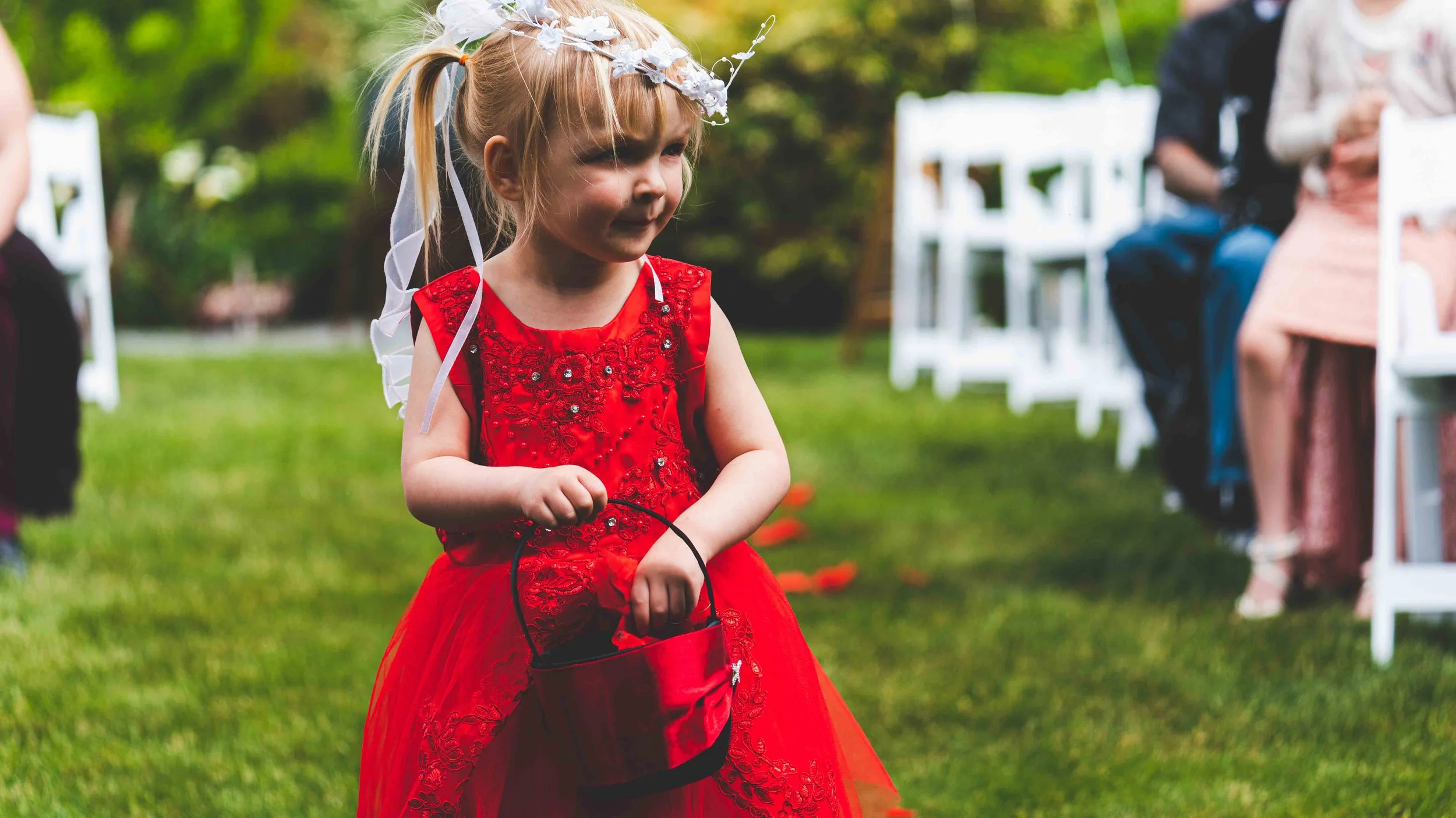 A young girl wearing a red dress with lace and sequins, holding a small red basket, at an outdoor event with white chairs and people in the background.