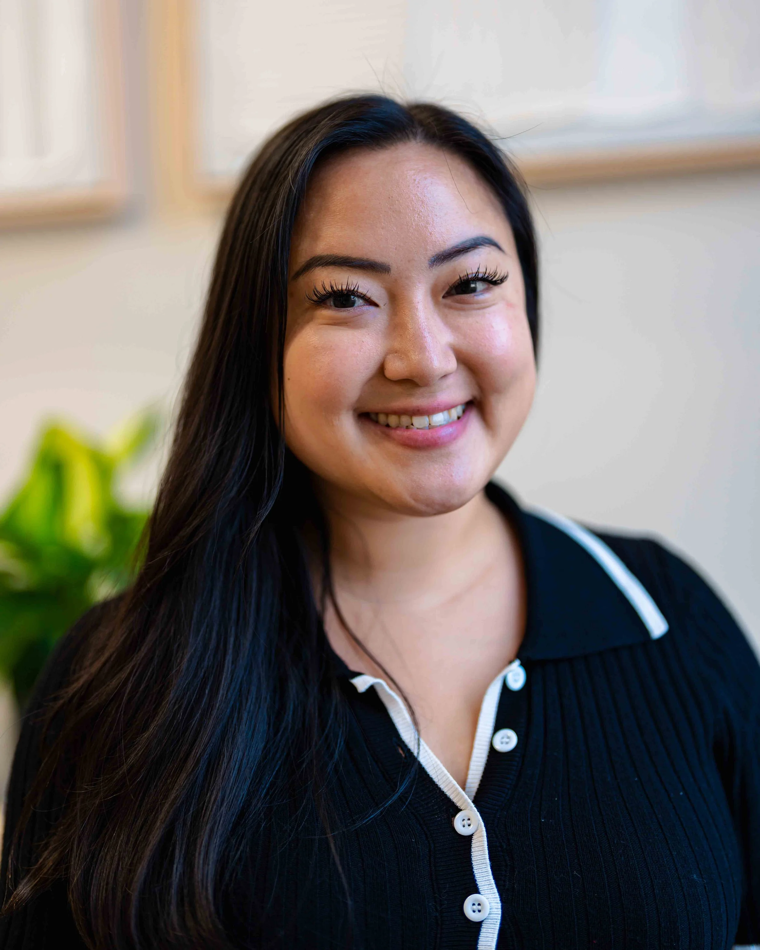 Close-up of a woman with long black hair, smiling, wearing a black shirt with white buttons and white trim.