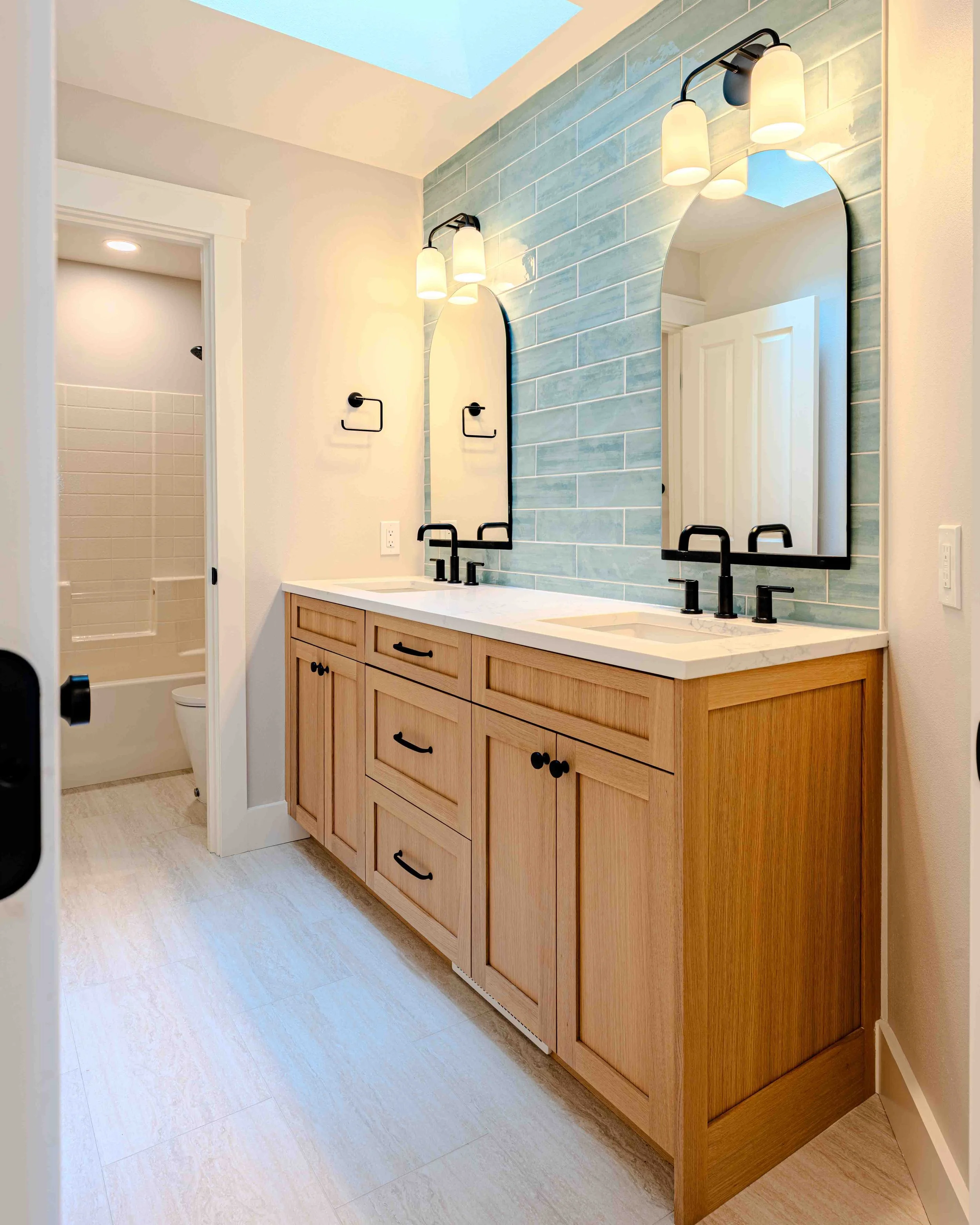 A modern bathroom vanity with a light wood cabinet and black hardware, a white countertop, double black faucets, and two arched mirrors. The wall behind the vanity features light blue rectangular tiles, and there are two wall-mounted light fixtures w