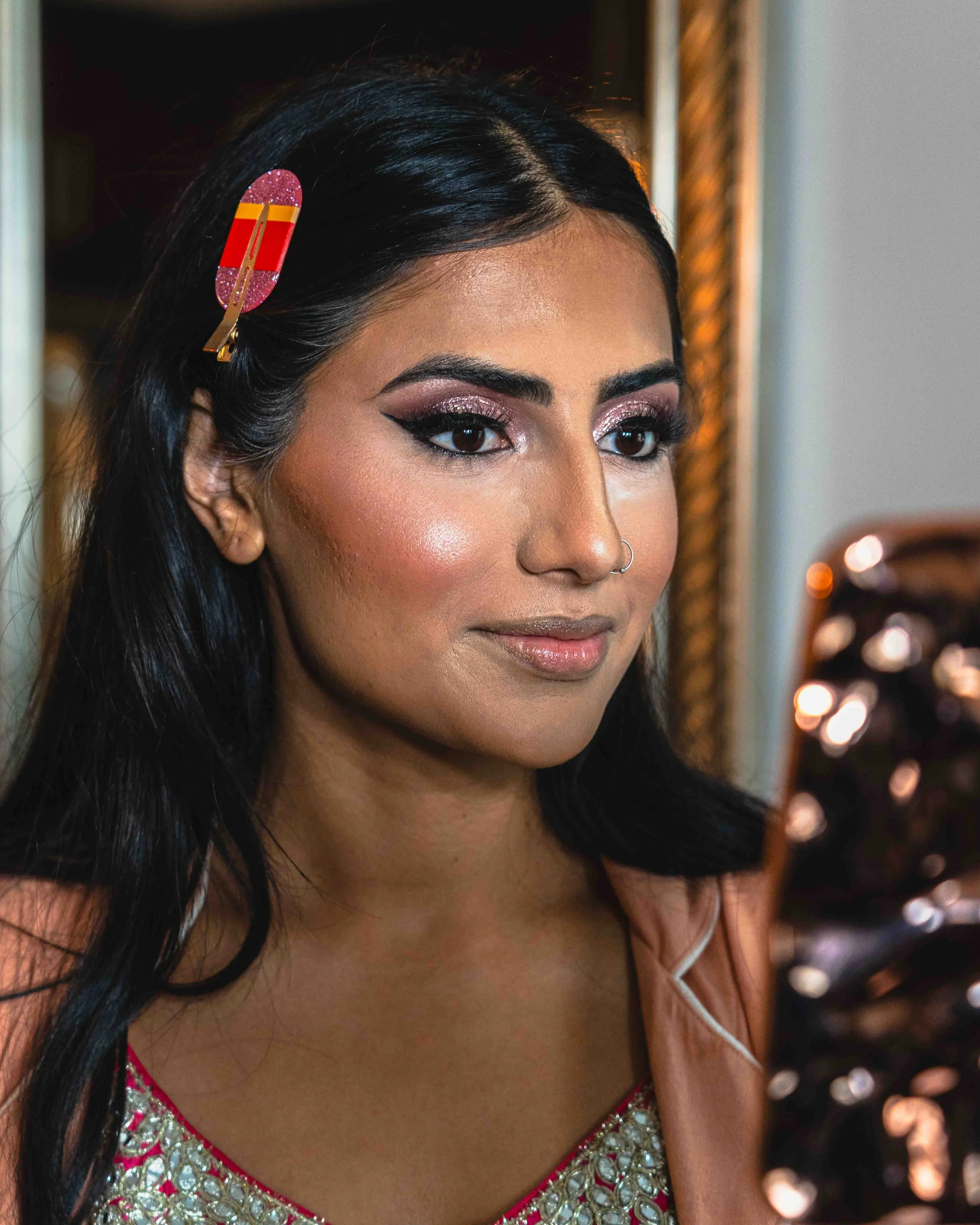 Close-up of a woman with dark hair, wearing a red and gold dress with intricate embellishments, and a pink glittery hair clip, against a backdrop with bokeh lights.