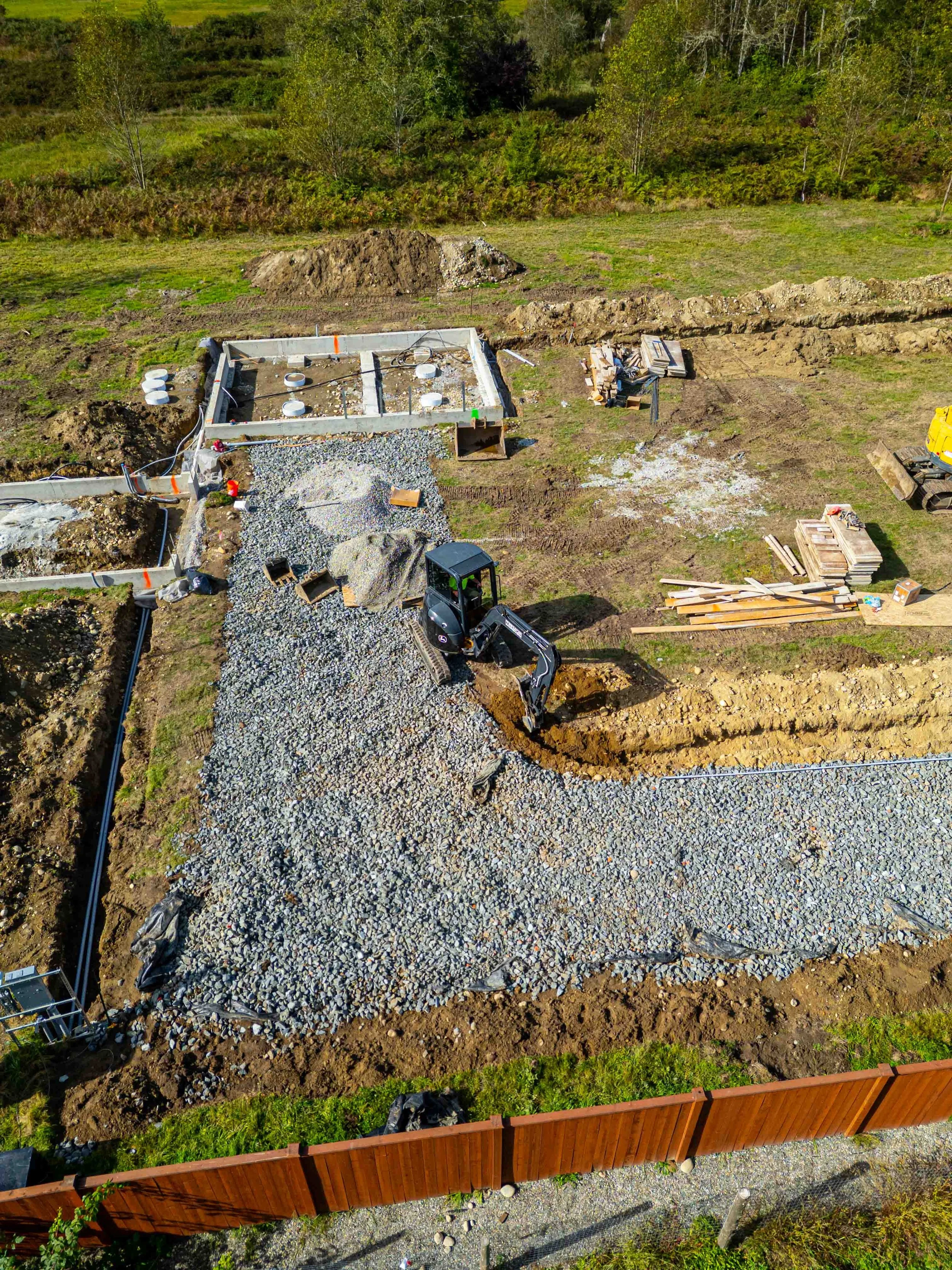 An aerial view of a construction site with foundation walls, gravel, construction materials, and machinery amidst green landscape and trees.