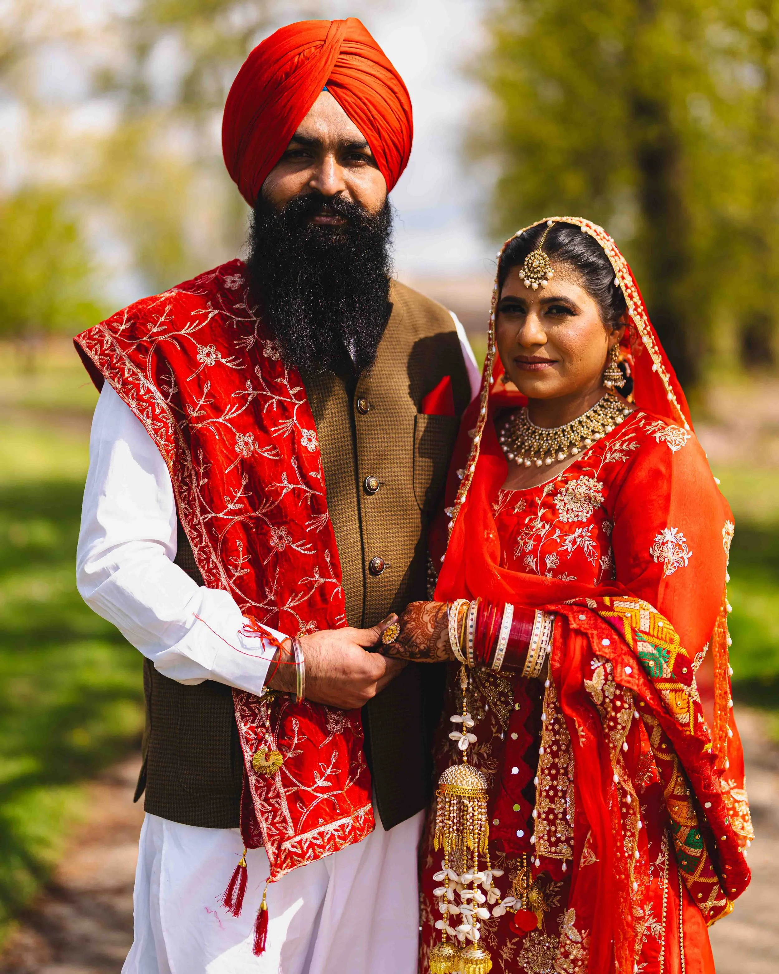 An Indian couple dressed in traditional wedding attire, the man wearing a white kurta with a red embroidered dupatta and the woman wearing a red saree with gold jewelry, standing outdoors.