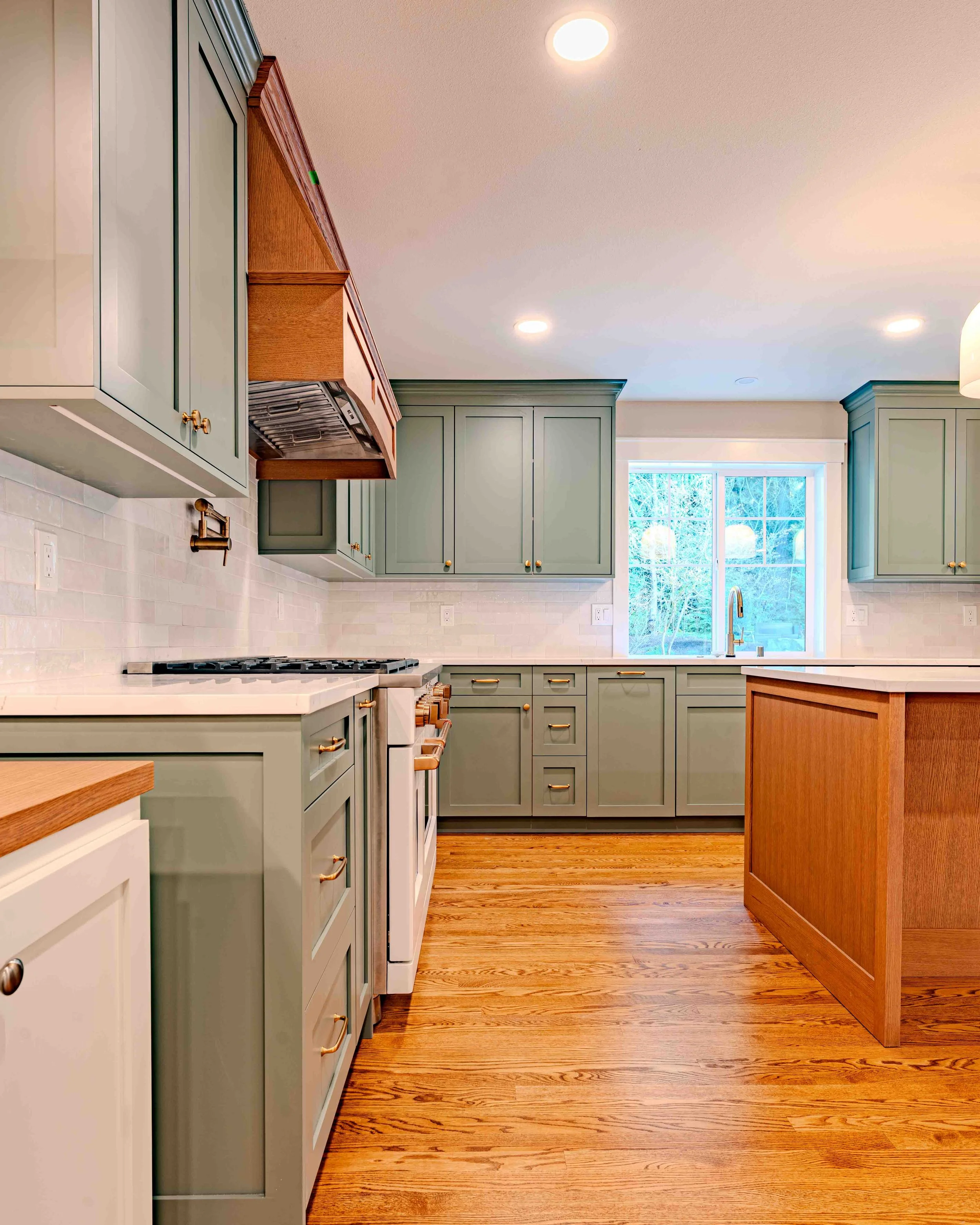 A modern kitchen with sage green cabinets, white countertops, a white stove, wooden flooring, a large window, and recessed ceiling lights.