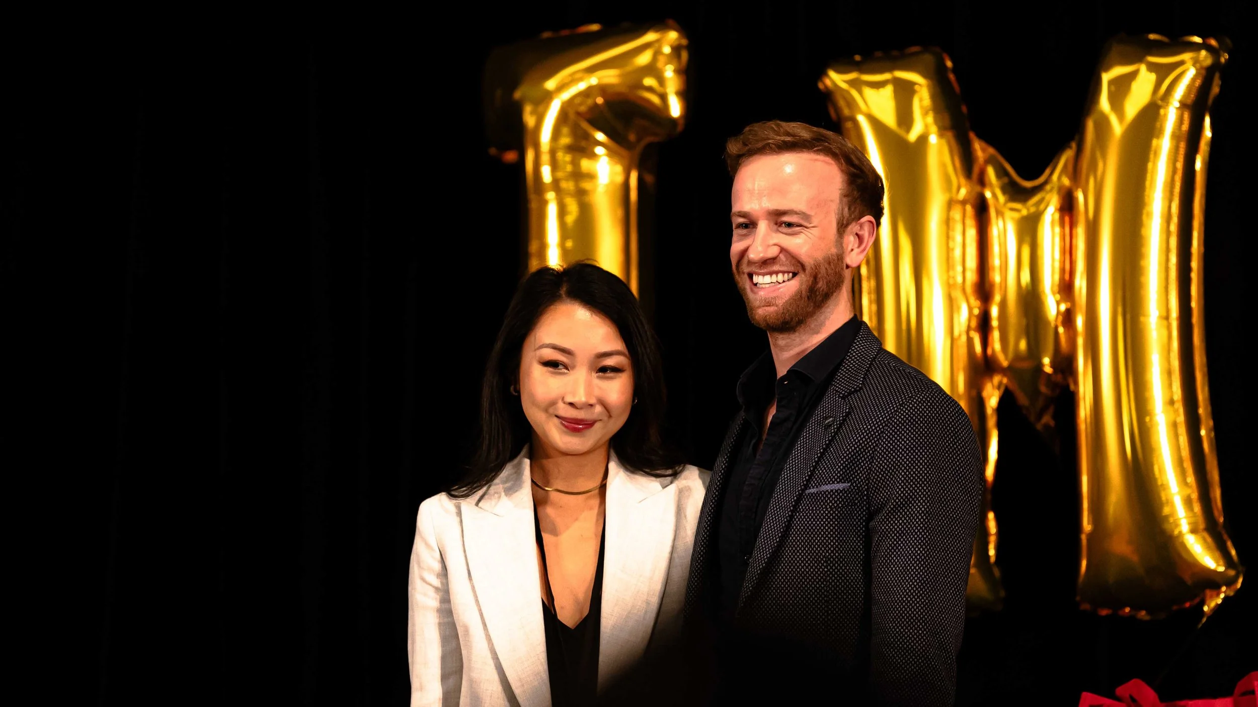 Two people, a woman and a man, smiling and standing close together at a celebratory event with golden balloons spelling out '2024' in the background.