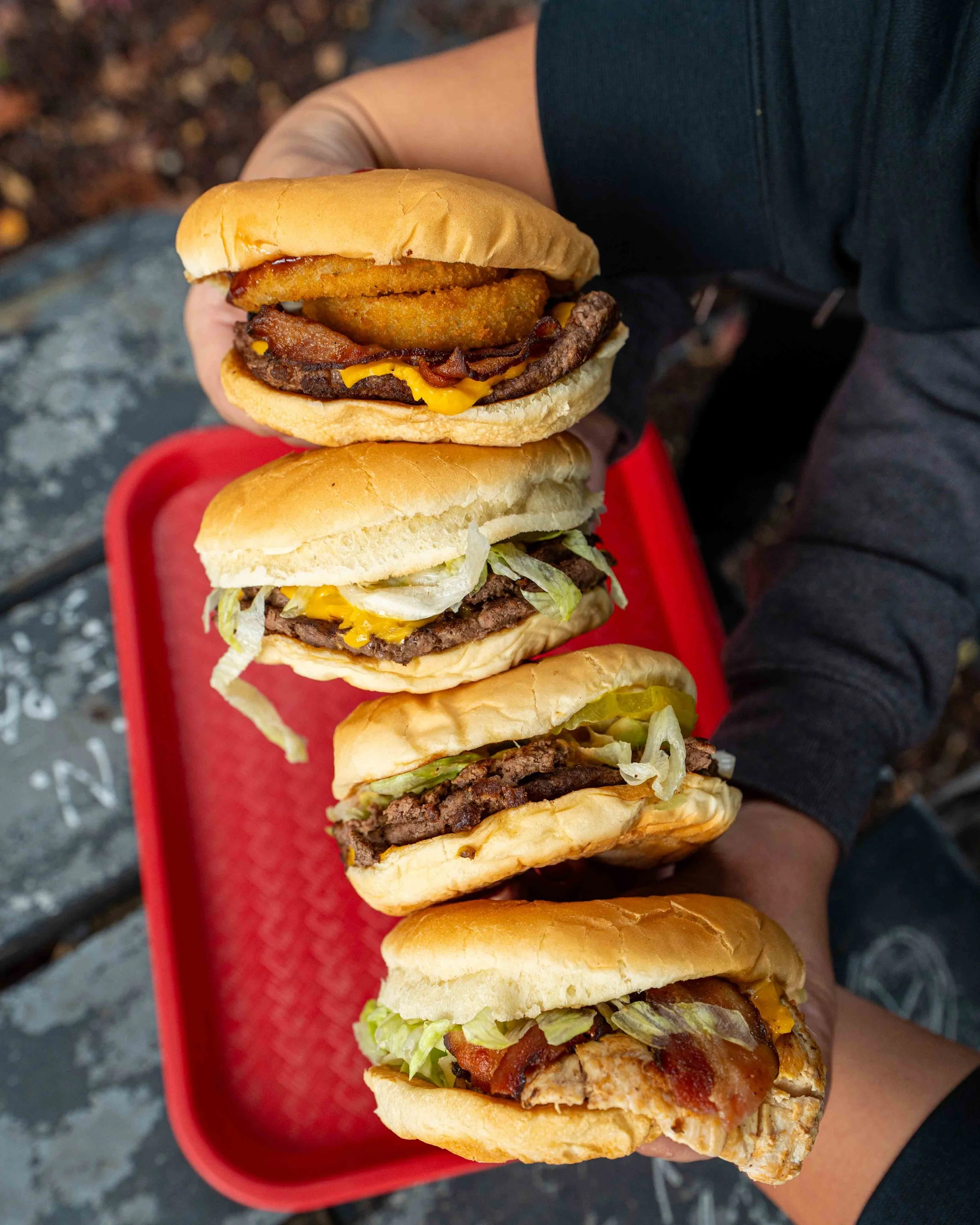 Close-up of a person holding a large sandwich with multiple layers including beef patty, lettuce, bacon, cheese, onion rings, and a bun, with a red tray underneath.