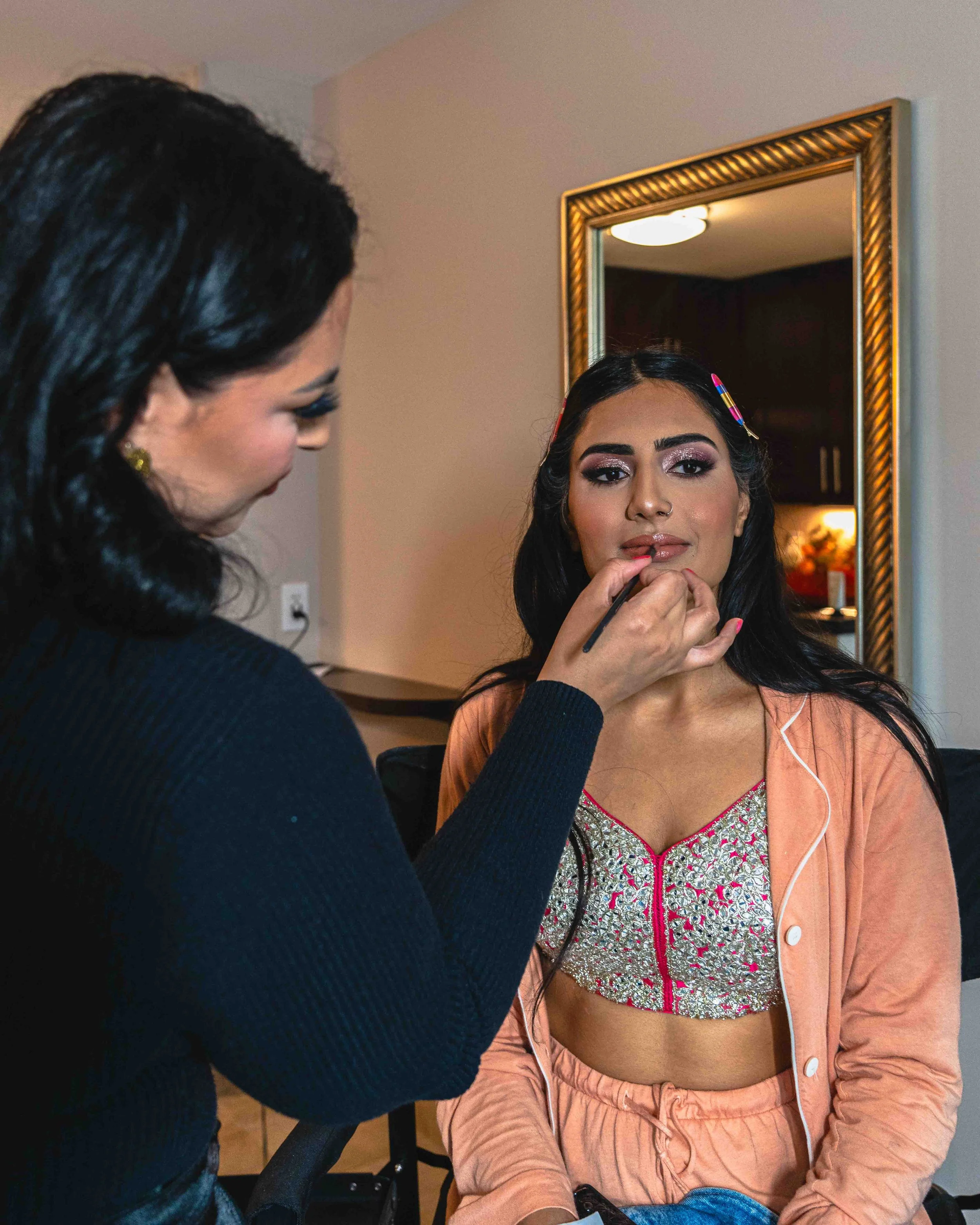 A makeup artist applying makeup to a woman sitting in front of a mirror in a room with a beige wall and warm lighting.