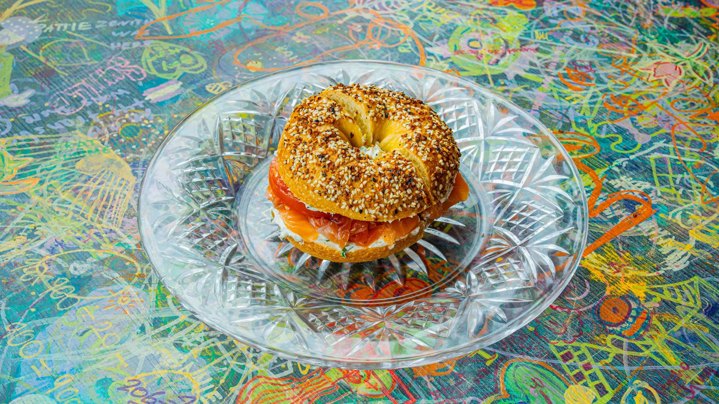 A bagel sandwich with smoked salmon, cream cheese, and tomato on a glass plate, placed on a colorful, graffiti-style tablecloth.