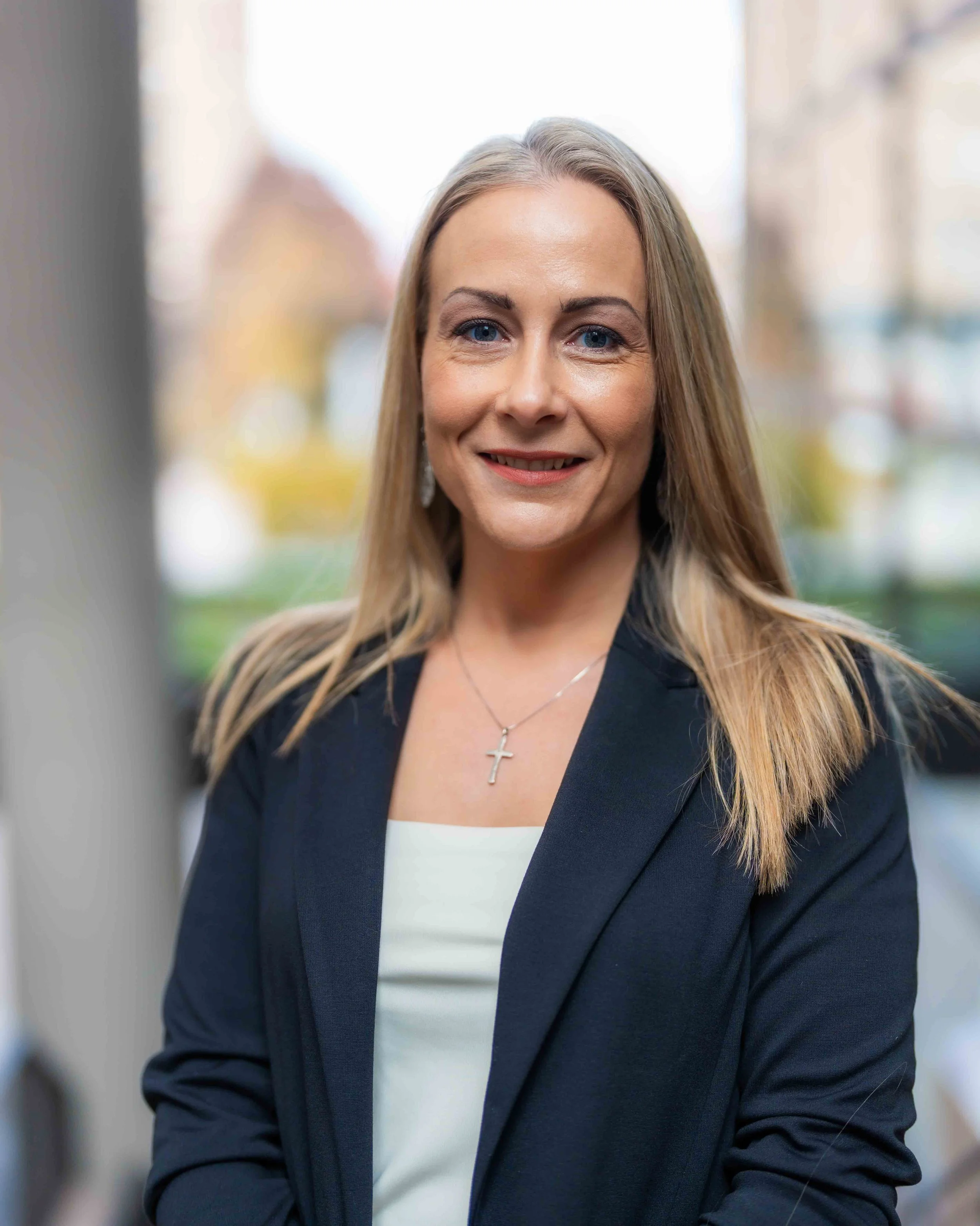 Close-up of a smiling woman with long blonde hair, blue eyes, wearing a black blazer over a white top, accessorized with a silver cross necklace, outdoors with blurred background of buildings and greenery.