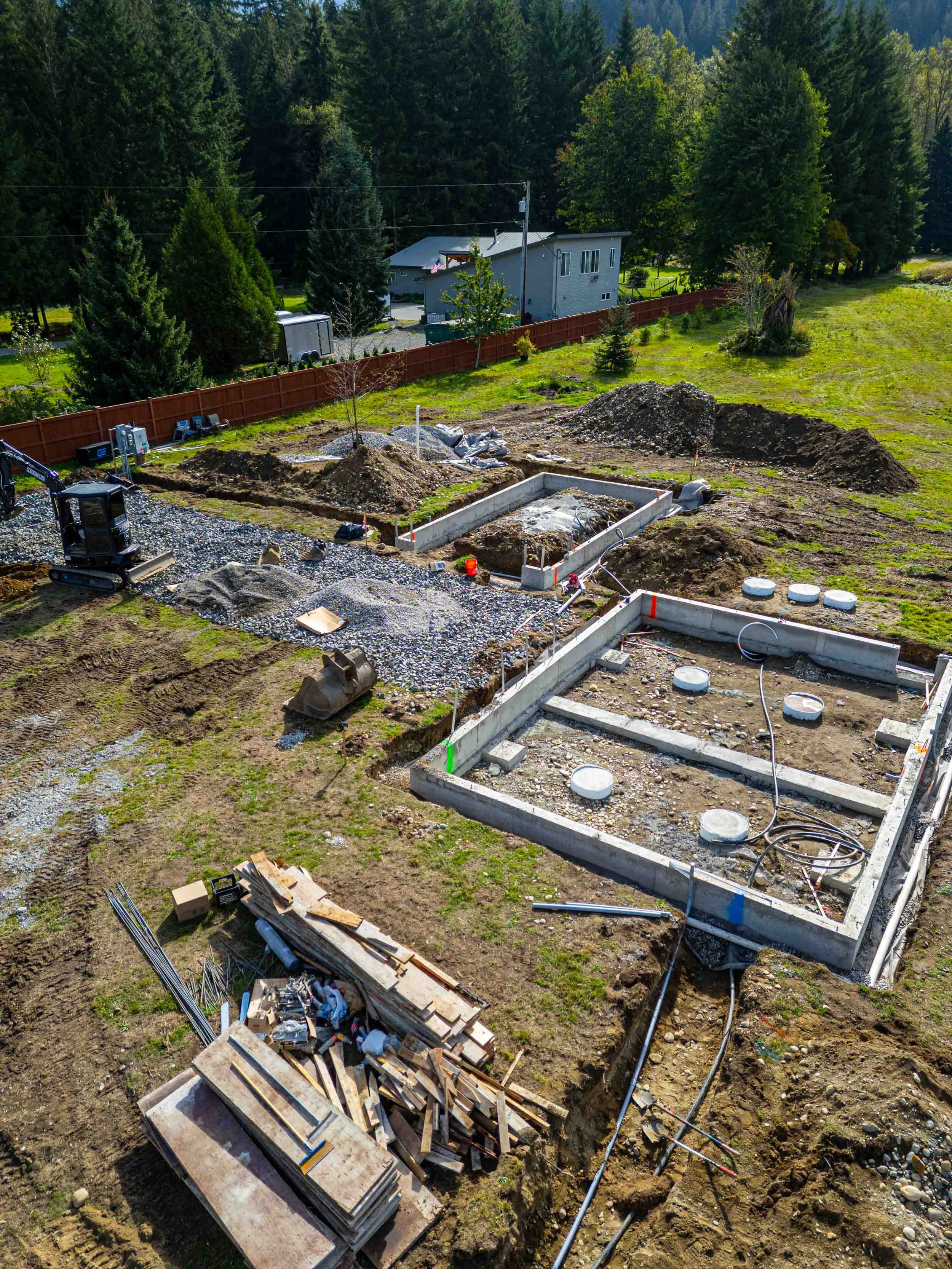 Construction site in a backyard with concrete footings, dirt, and building materials, surrounded by green grass and trees.