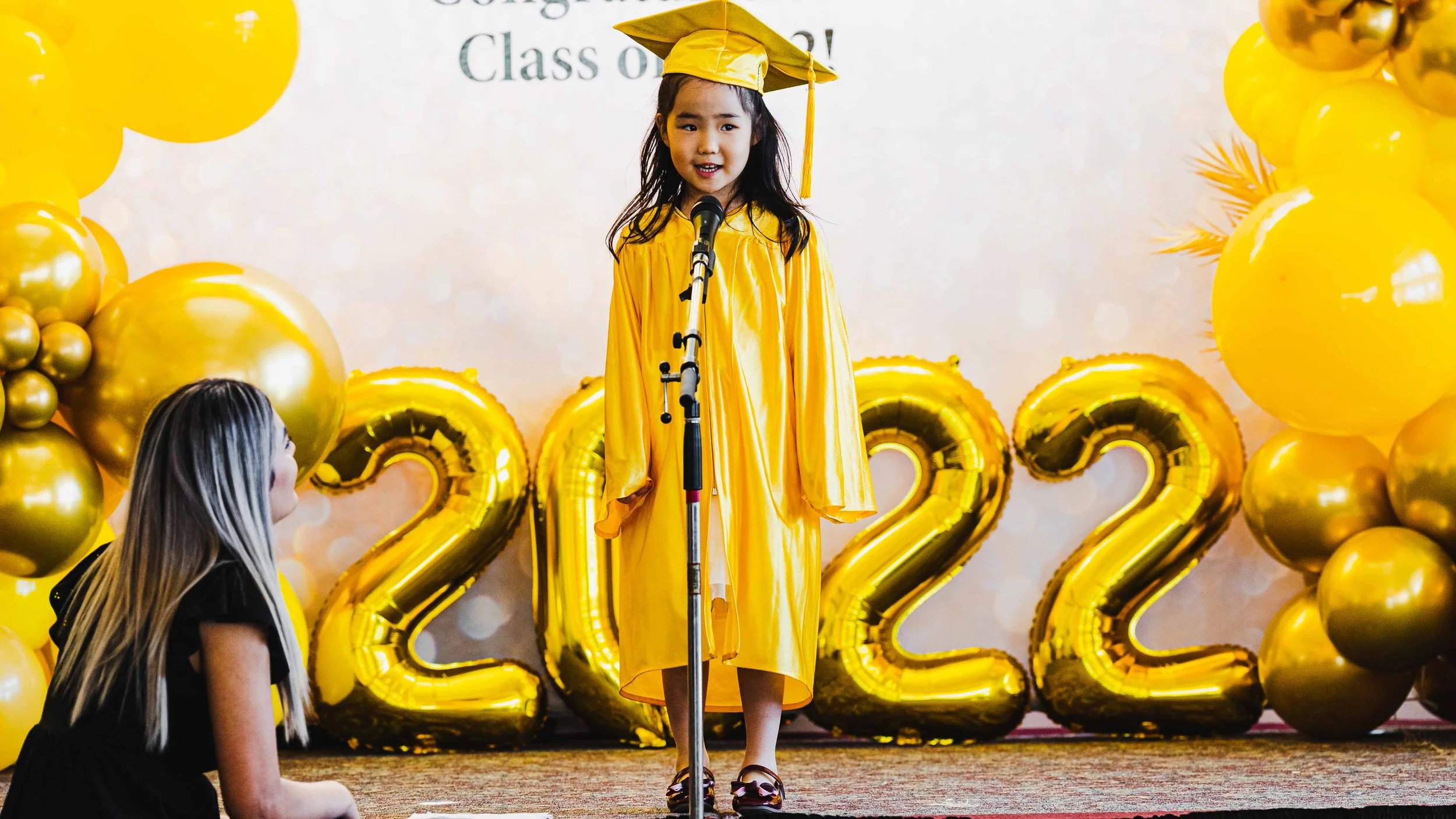 Young girl in yellow graduation cap and gown speaking at a microphone during a graduation ceremony, with gold balloons and '2022' balloons in the background.