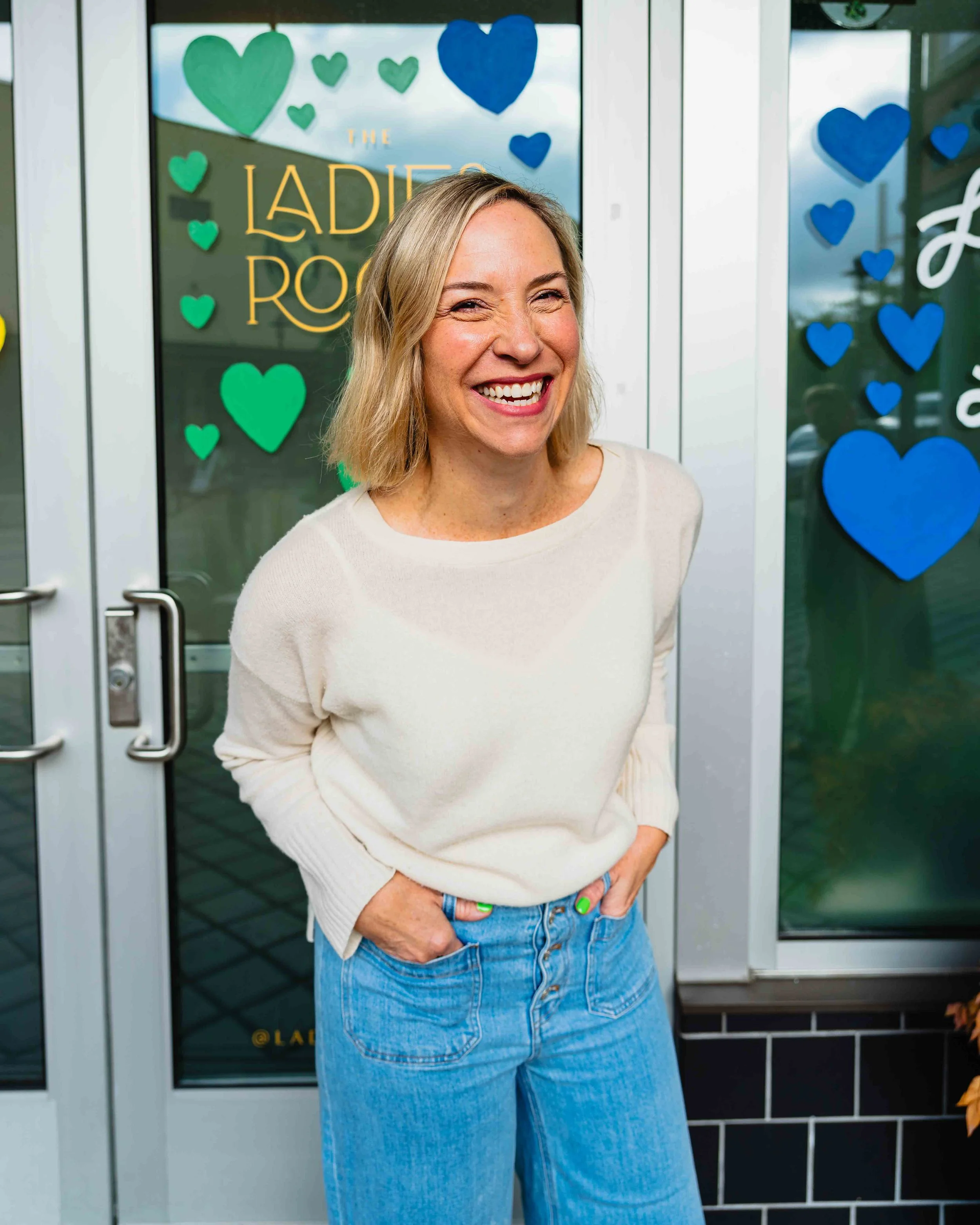 A woman smiling with her eyes squinting, standing in front of a door decorated with blue and green paper hearts, with a sign reading 'The Ladies Room' in yellow lettering.