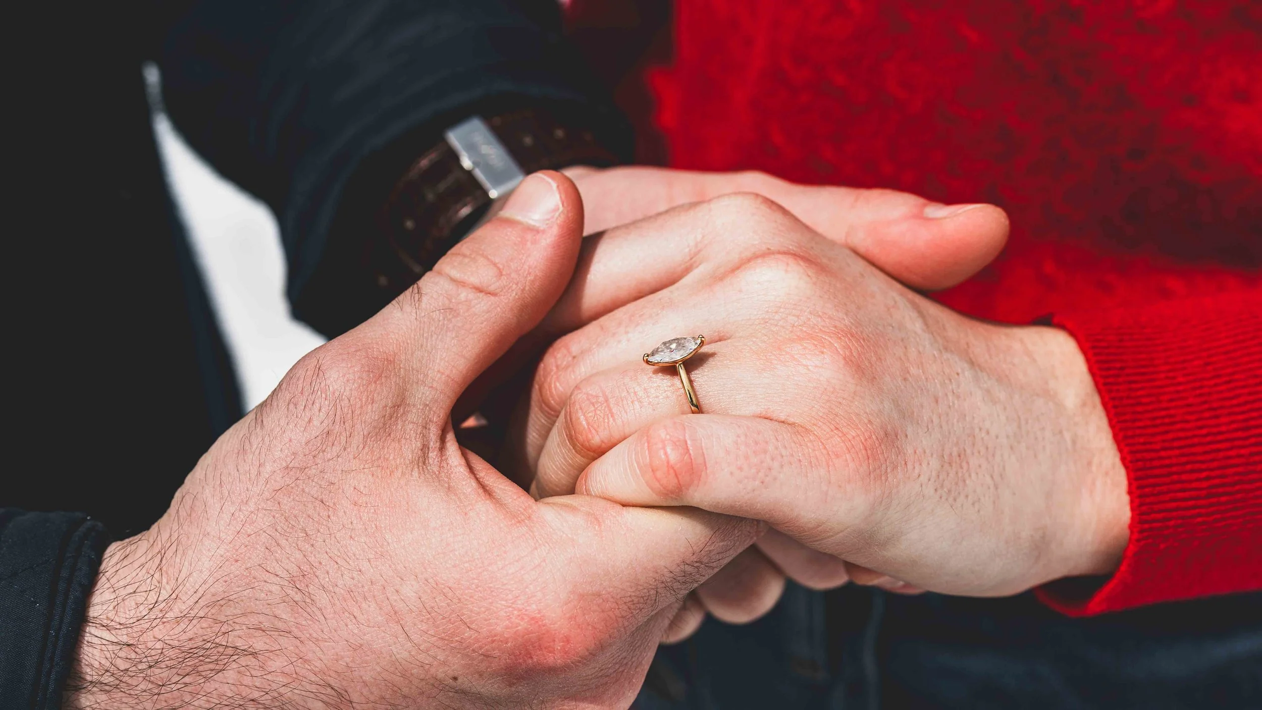 A close-up of two people holding hands, with one person wearing a red sweater and a diamond ring visible on their finger, and the other person wearing a black long-sleeve shirt or jacket.