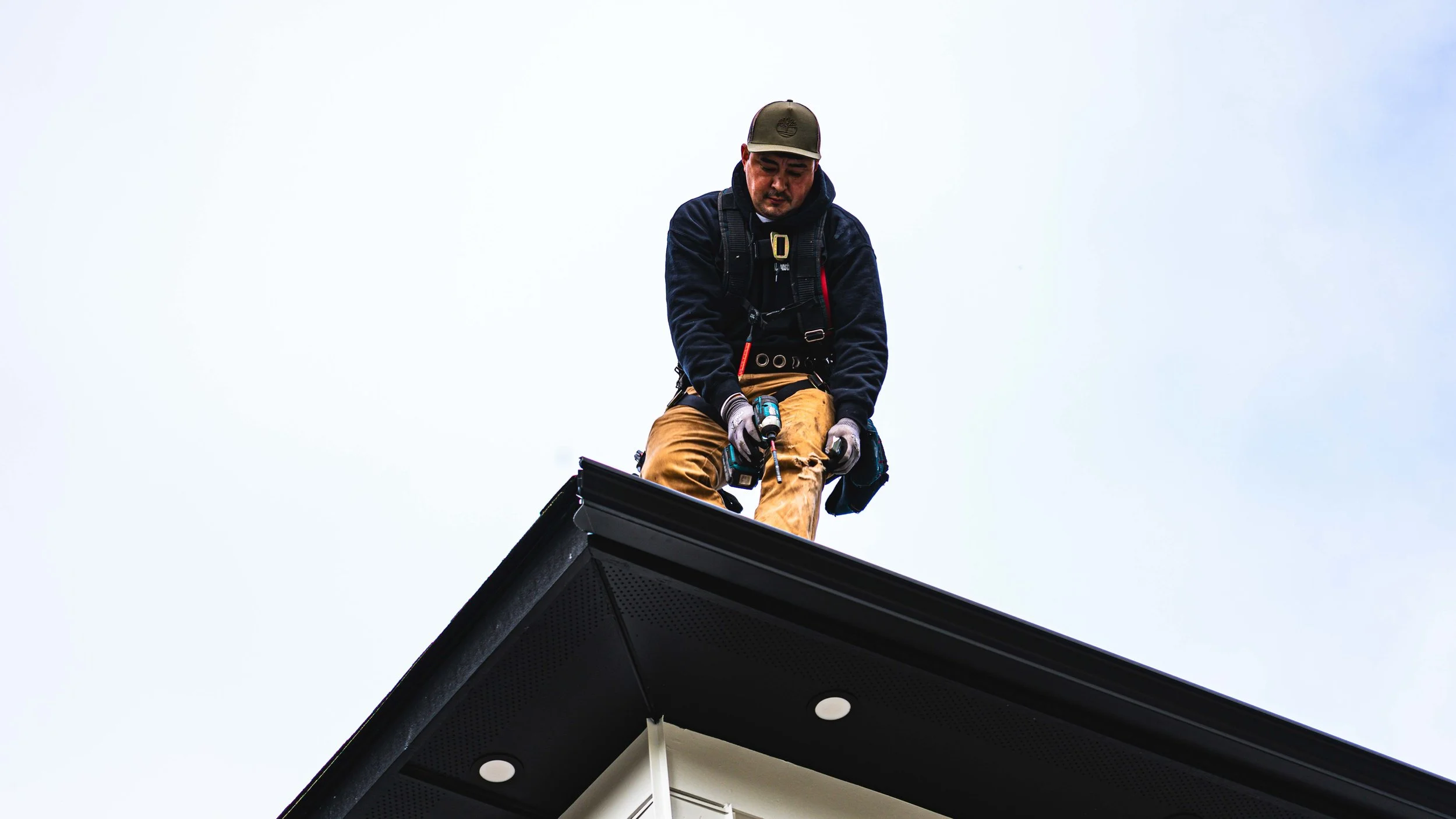A man wearing a cap, black hoodie, and brown pants is working on the roof of a house with the sky in the background.