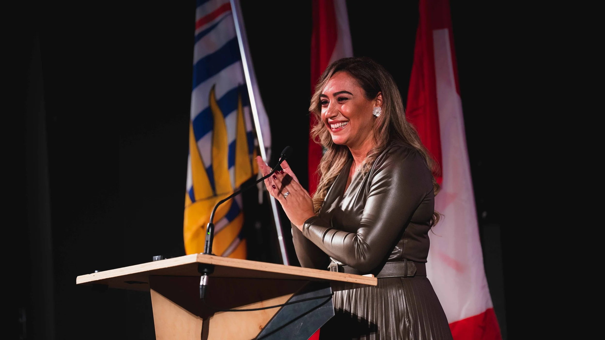 A woman with long wavy hair, wearing earrings and a shiny brown jacket, is smiling and speaking at a podium with a microphone, with flags in the background.
