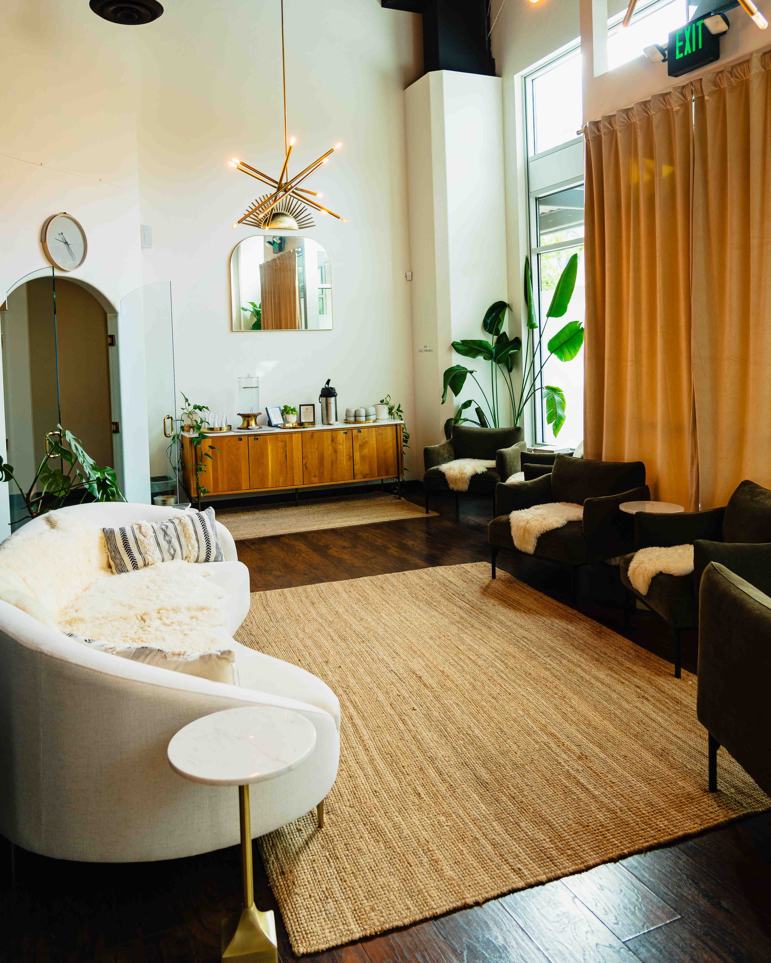 Interior of a modern lounge with a white sofa, black armchairs with fur cushions, a wooden sideboard with decorative items, large green plants, and beige curtains by a tall window. A circular wall clock and a contemporary chandelier are visible, with