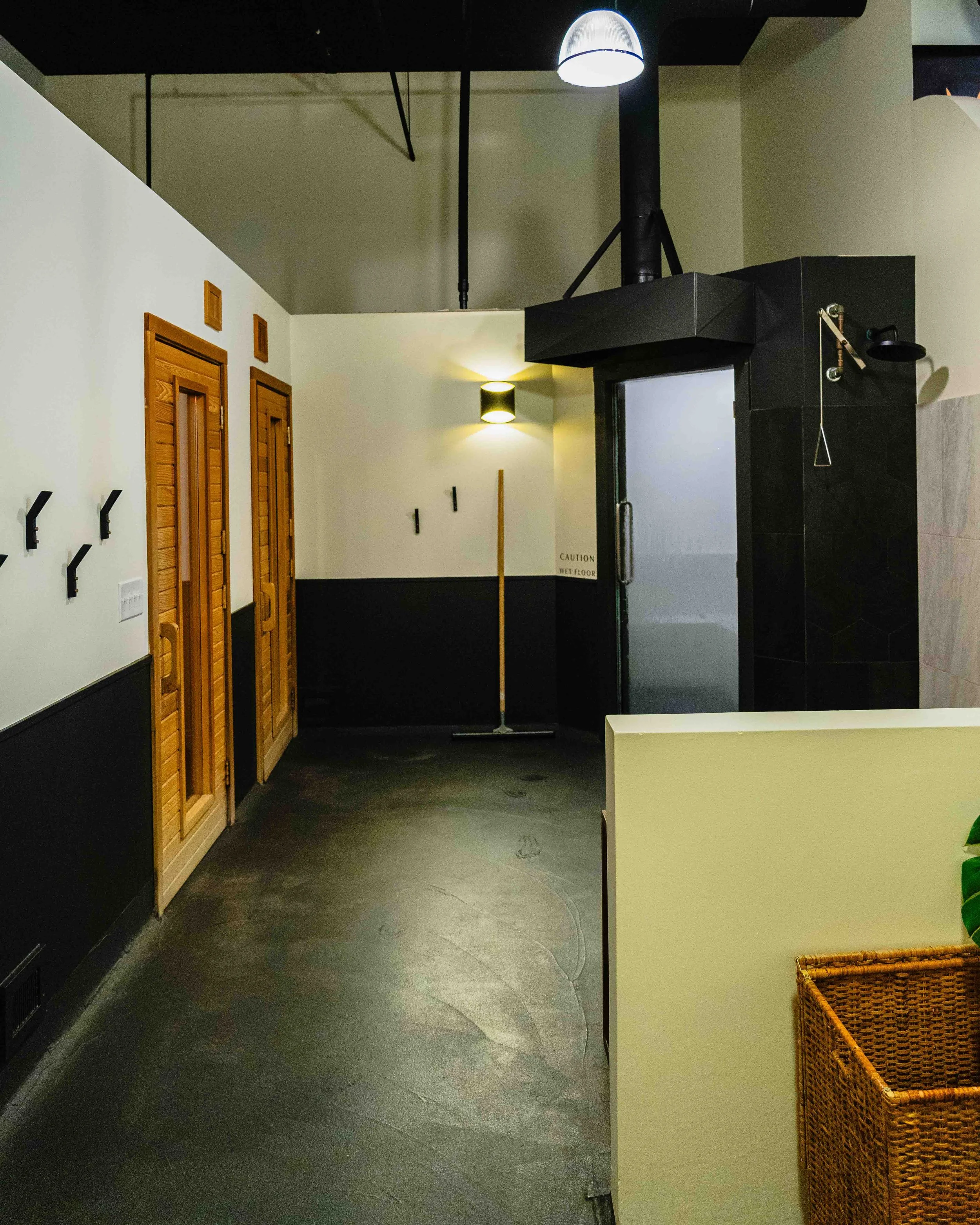Interior of a modern room with black flooring, white walls, wooden doors, and a black ceiling. There are coat hooks on the wall and a woven basket on the right. A yellowish light fixture is mounted on the wall, and various ceiling fixtures are visibl