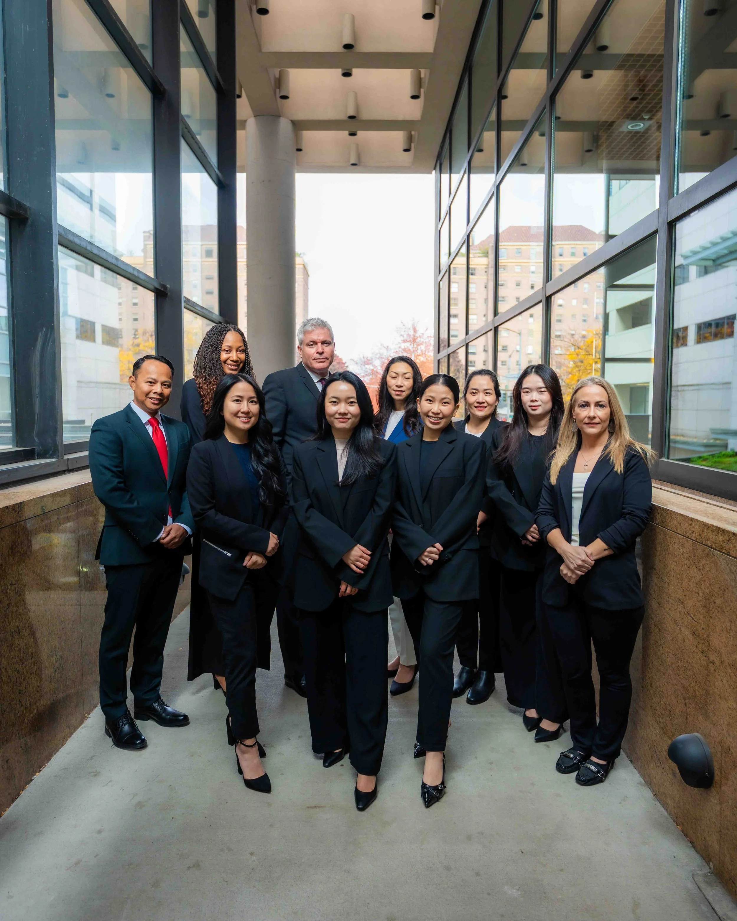 A group of professional men and women dressed in business attire posing together outdoors in a modern urban setting with glass buildings and cityscape in the background.
