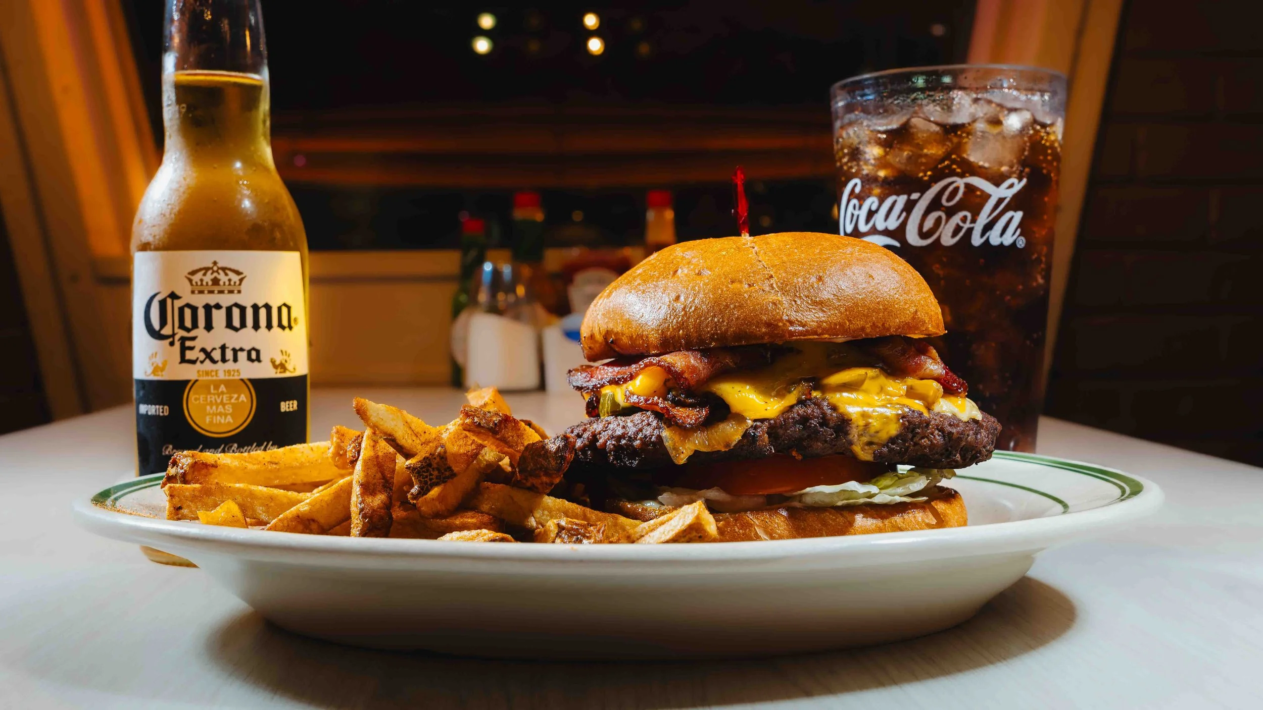 Plate with cheeseburger, French fries, and a drink with ice. In the background, a bottle of Corona Extra beer and a glass of Coca-Cola.