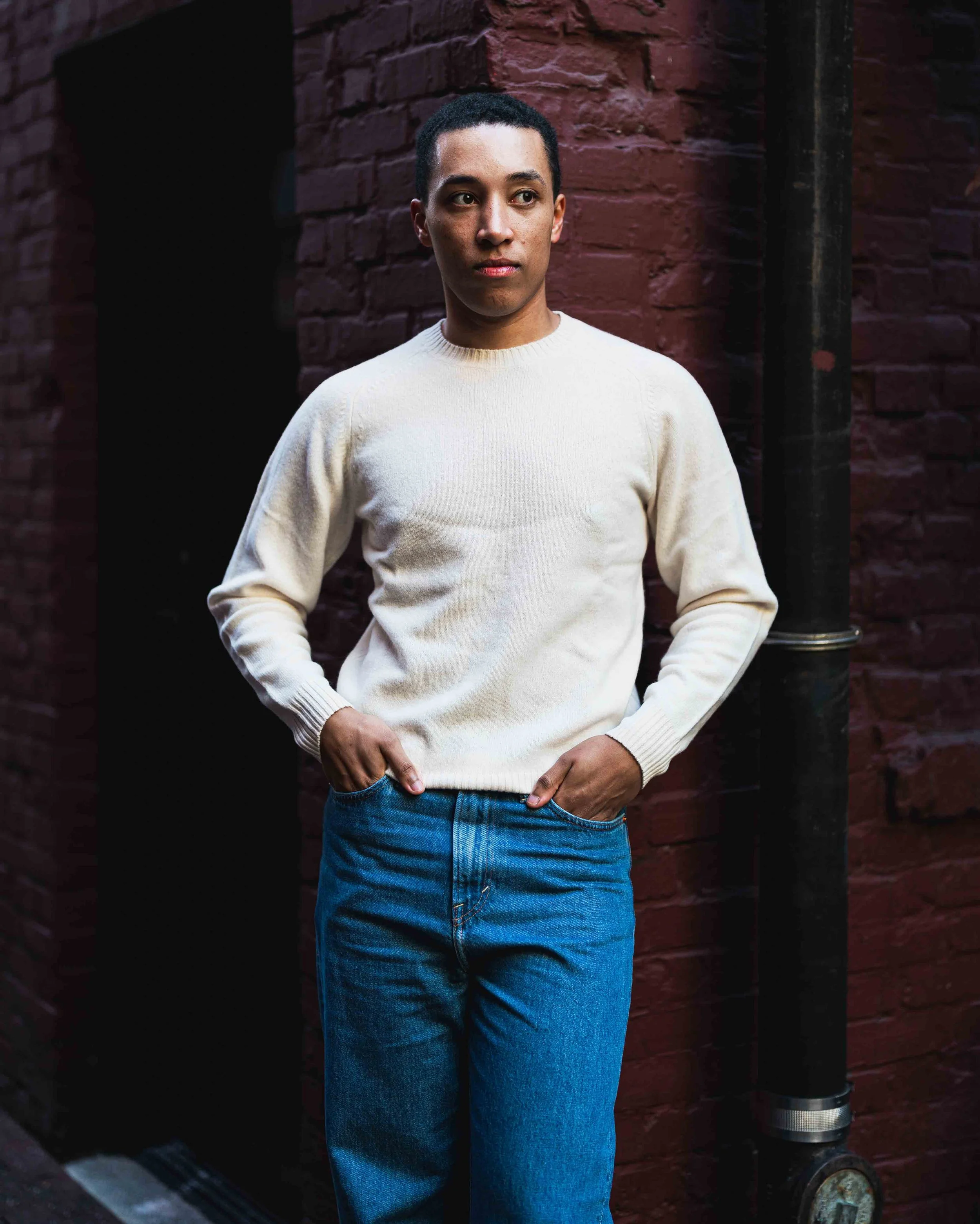 A person with short black hair and medium skin tone standing with hands on hips, wearing a white sweater and blue jeans, against a red brick wall with a black pipe and a fire hydrant valve.