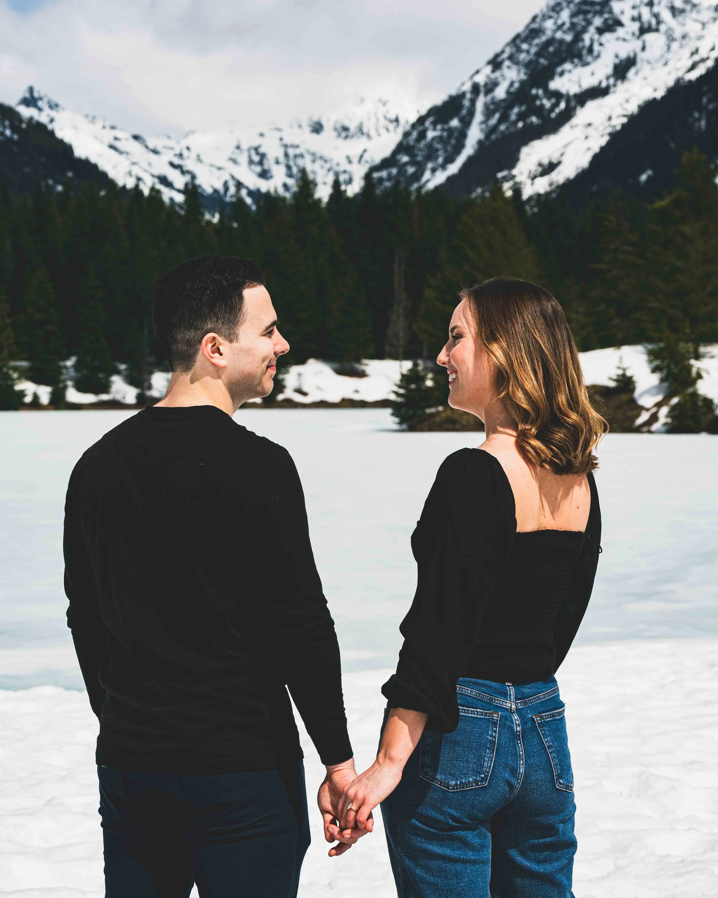 A couple standing on snow-covered ground, holding hands, facing each other with smiles, with snow-capped mountains and a forest of evergreen trees in the background.