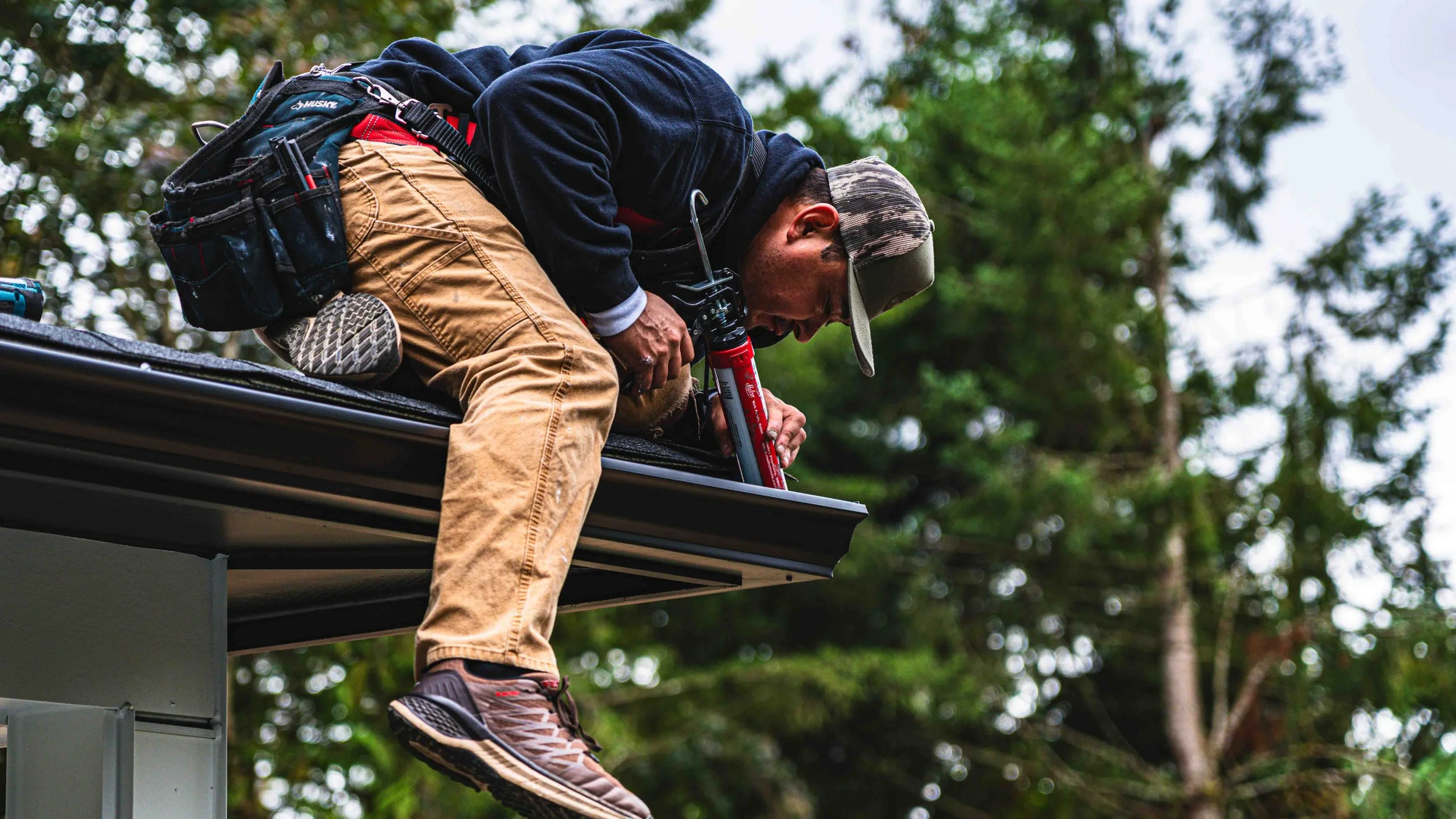 A man kneeling on a roof, installing or repairing a gutter, surrounded by trees and outdoor scenery.