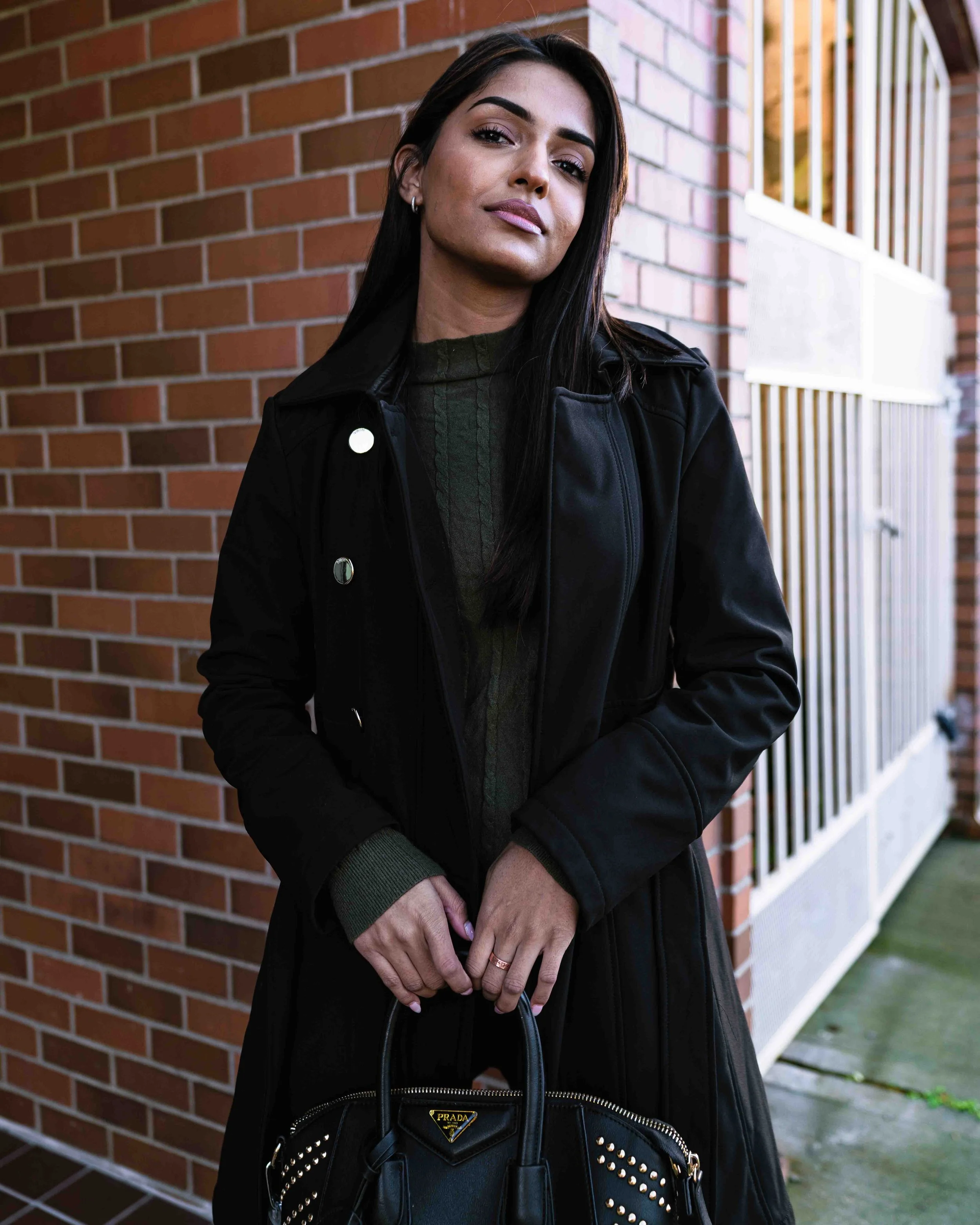 A woman with long dark hair and makeup, dressed in a black leather jacket, holding a black Prada handbag with gold studs, standing against a brick wall.