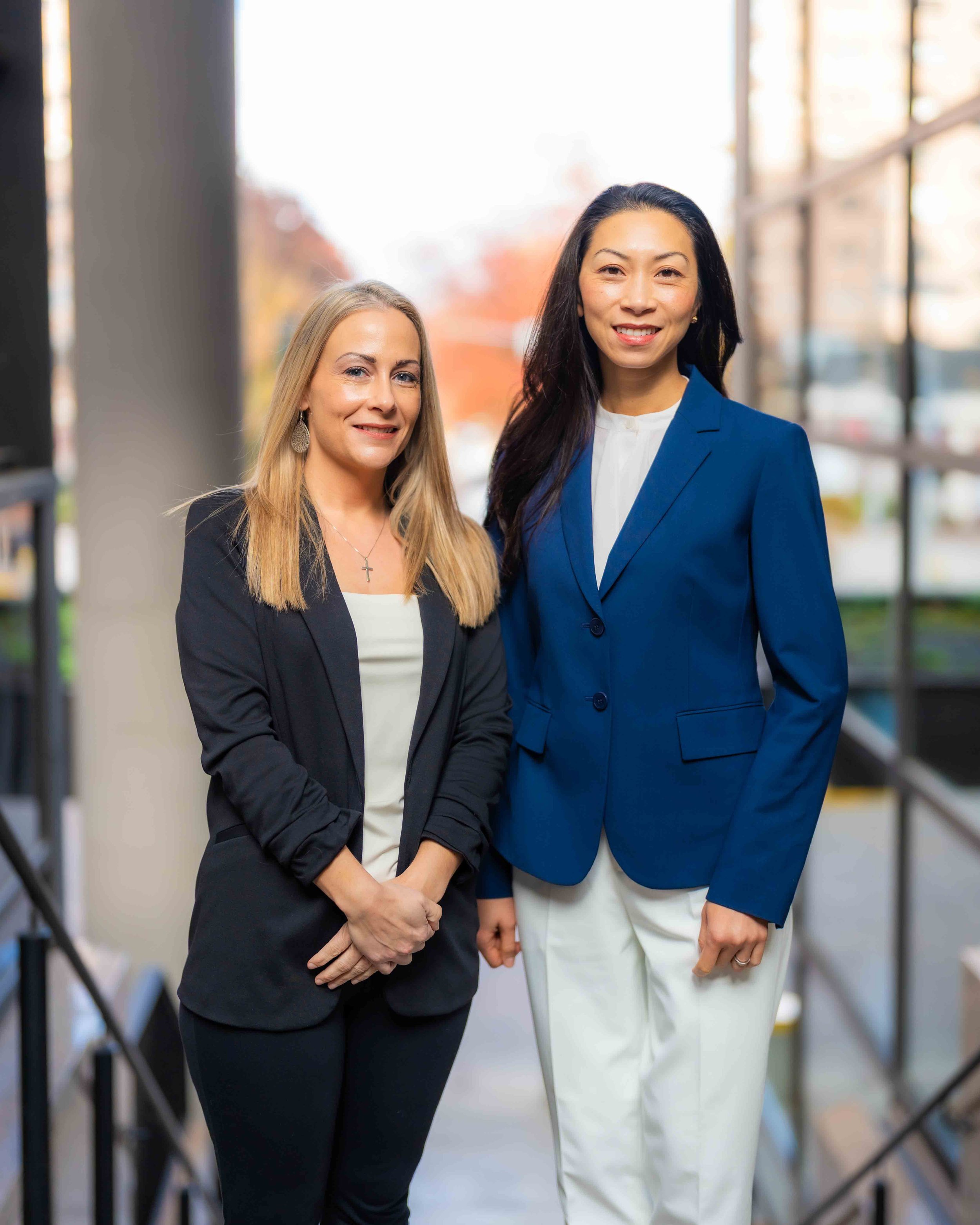 Two women standing close together outdoors in fall, smiling at the camera, holding hands, with a blurred background of a building and trees.