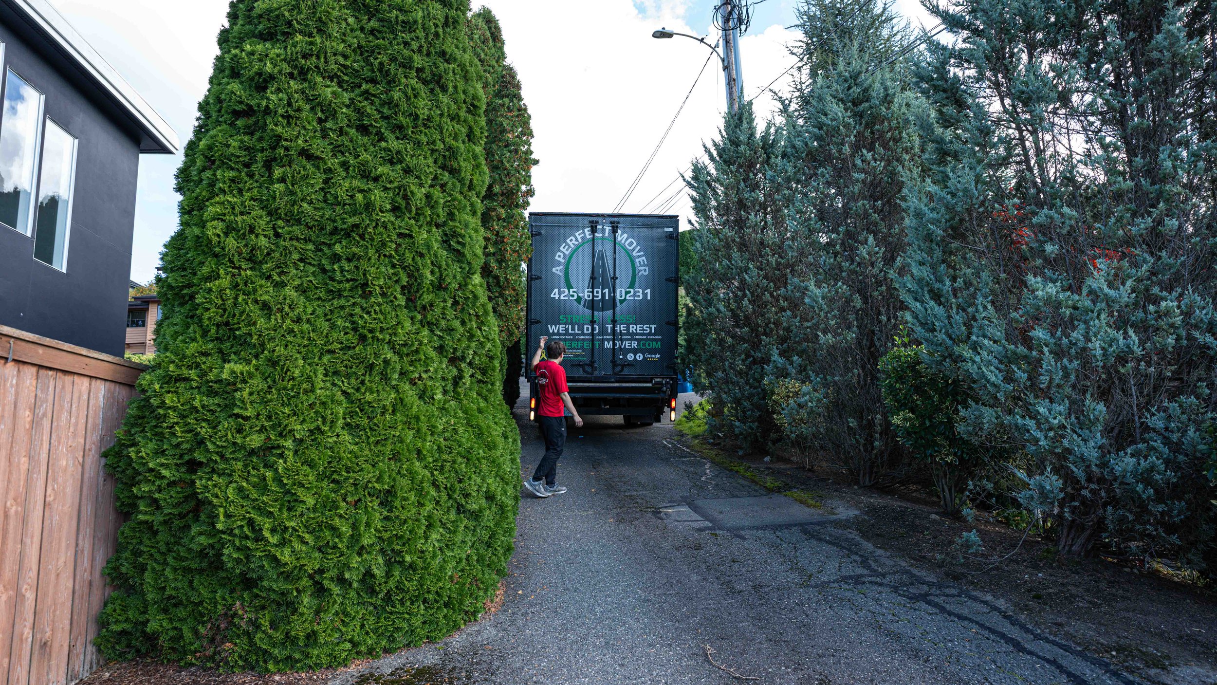 A person in a red shirt standing next to a truck with a sign that reads 'A Perfect Mover,' parked on a narrow driveway between tall green trees and a black house with a wooden fence.