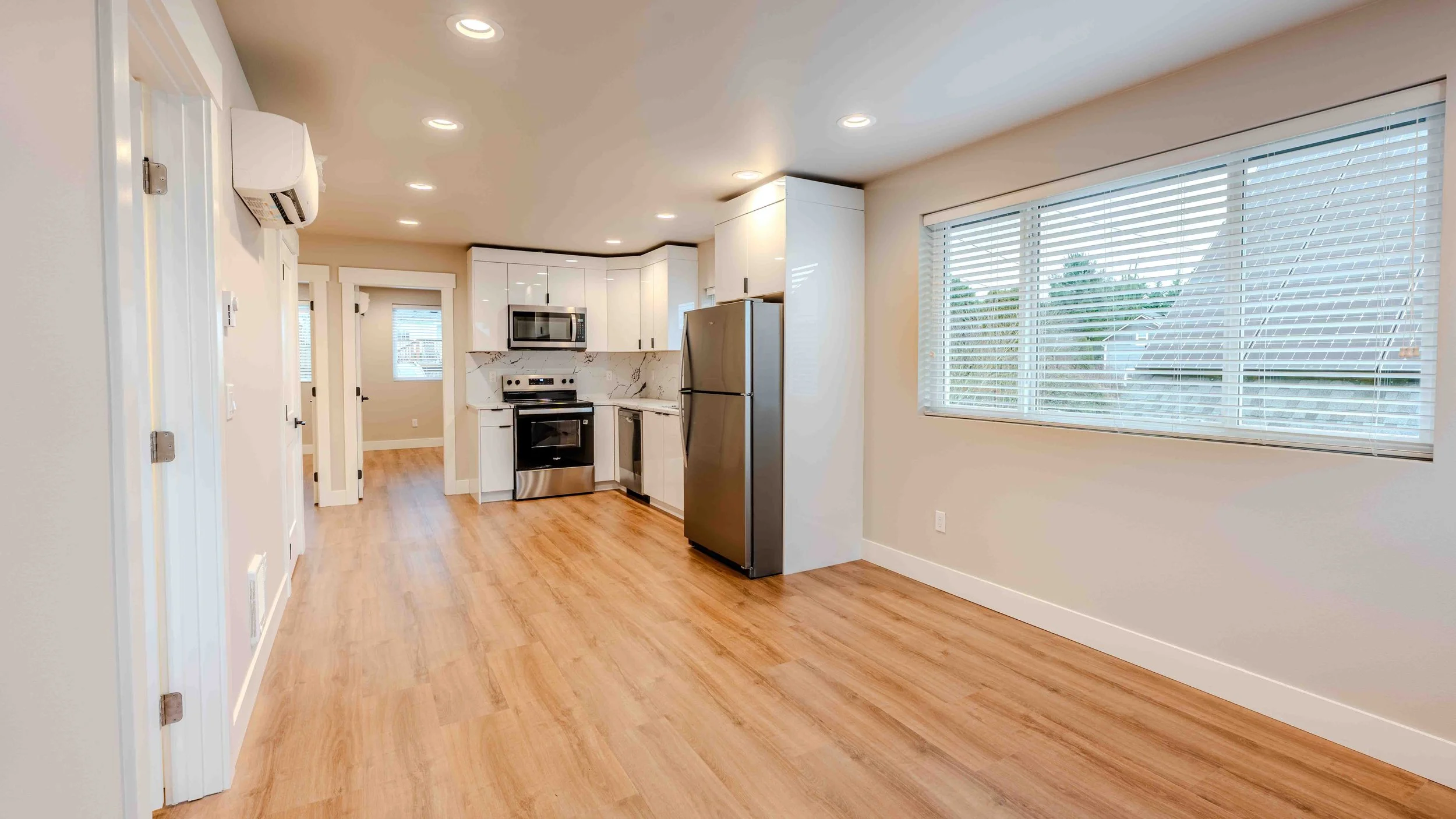 Interior view of a modern kitchen with white cabinets, stainless steel appliances, large window with blinds, and wooden flooring.