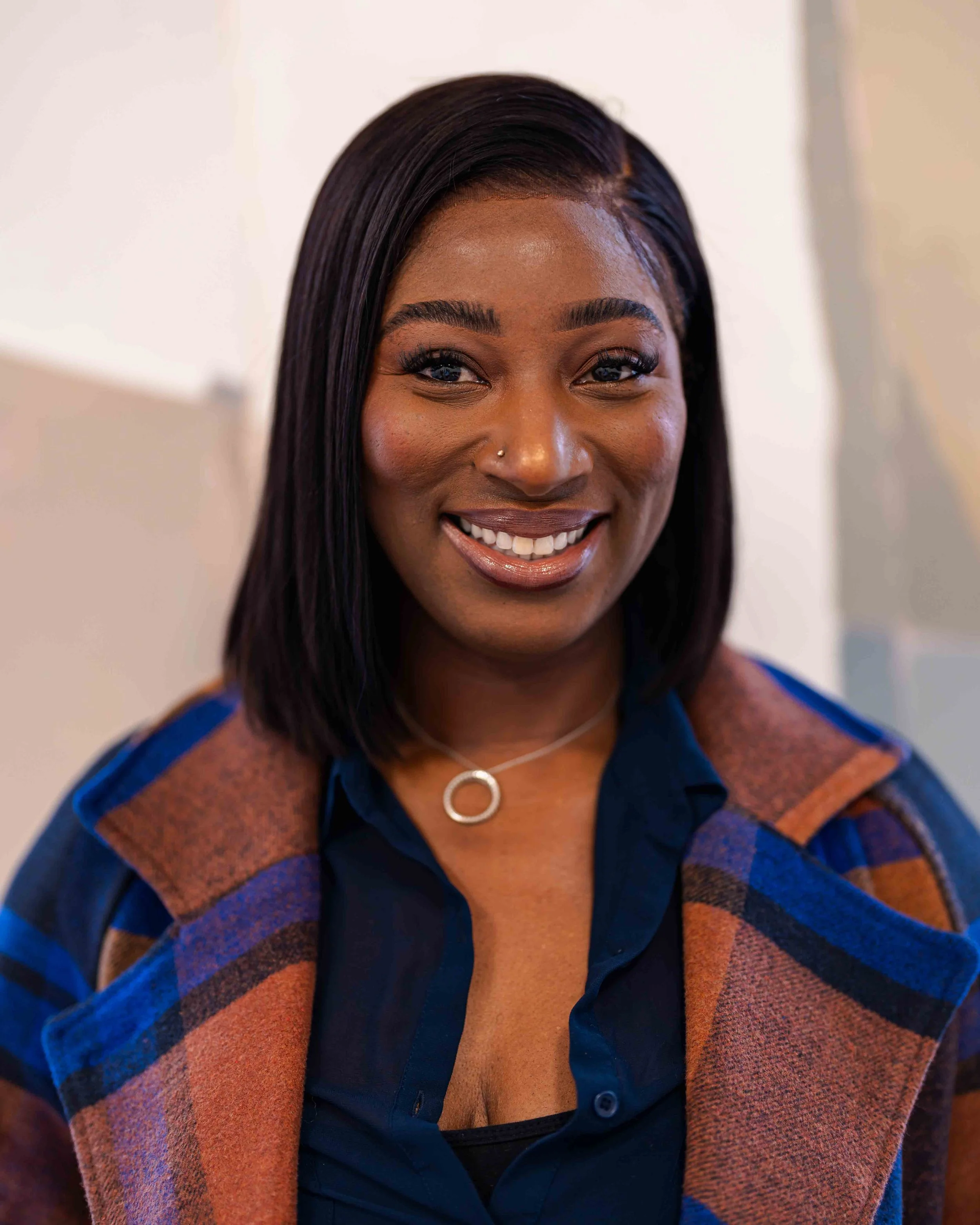 Close-up of a woman with dark hair, wearing a colorful plaid blazer and a navy blue shirt, smiling with visible teeth, in an indoor setting.
