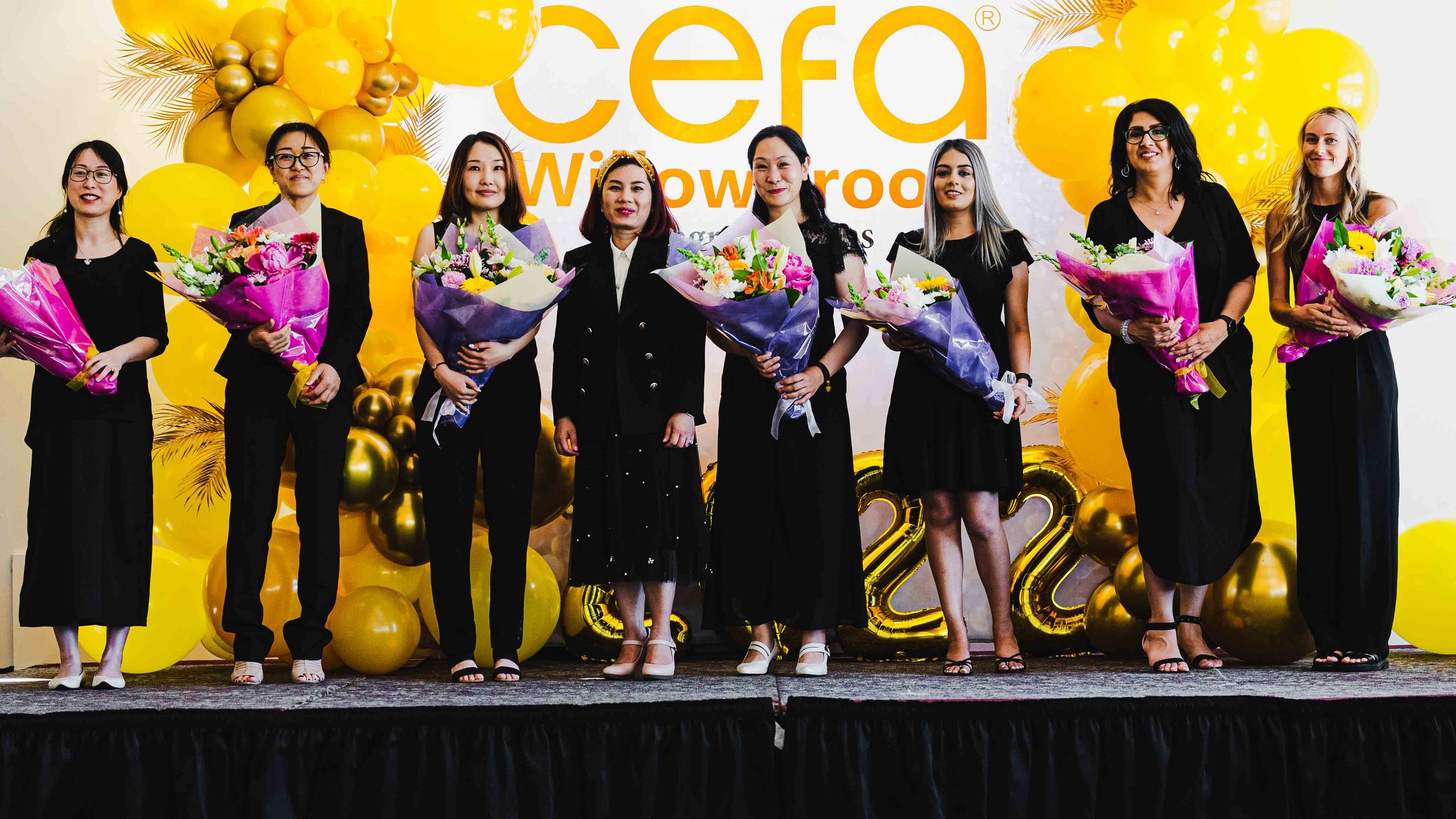 Women standing on stage with bouquets of flowers in front of a yellow backdrop that reads 'CEO' and 'Wow' with decorative balloons, celebrating a professional achievement or event.