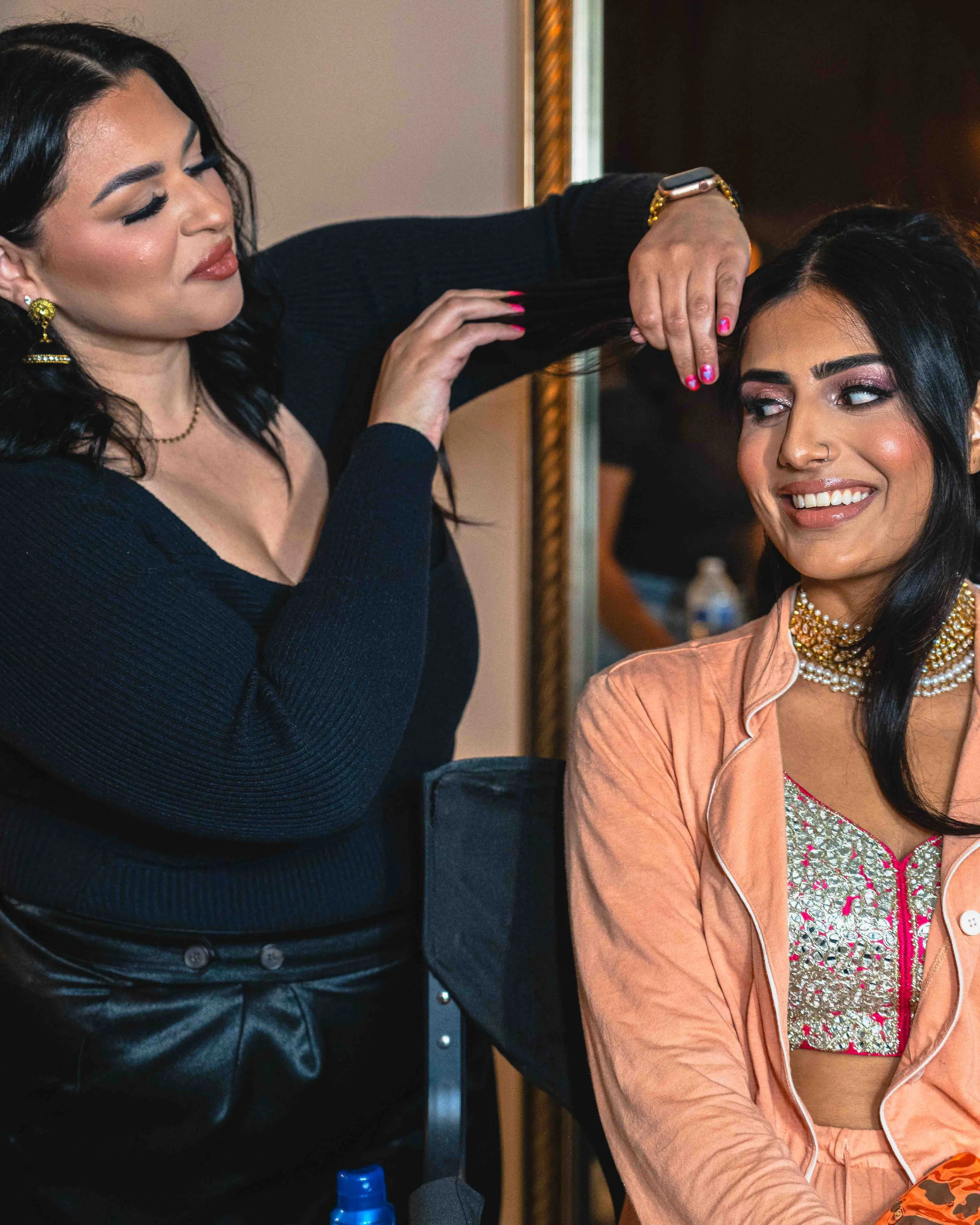 A woman with long dark hair is getting her hair styled by another woman. The hairstylist, wearing a black top, is working on the client's hair. The client is smiling, wearing a pinkish-orange jacket and a patterned blouse, with makeup and jewelry.