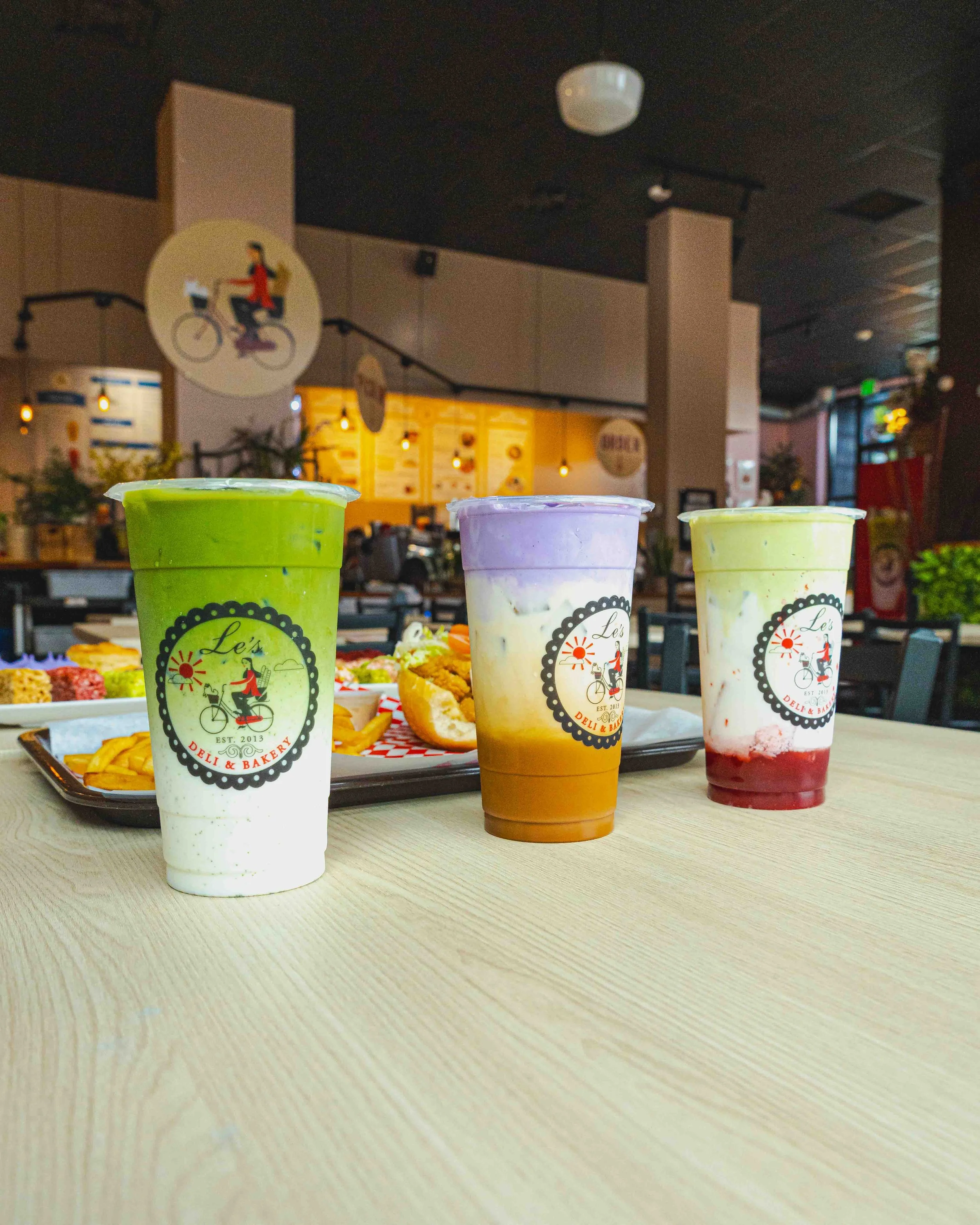 Three colorful beverages on a table, with a tray of assorted snacks and a blurry background of a restaurant or cafe interior.